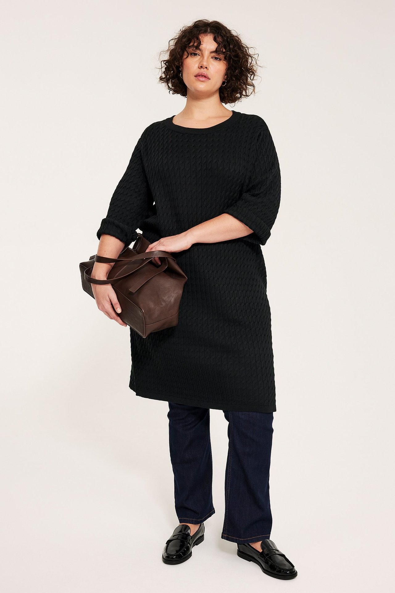 Framifrån, kvinna i svart kabelstickad klänning, mörkblå jeans, svarta loafers, håller brun handväska.