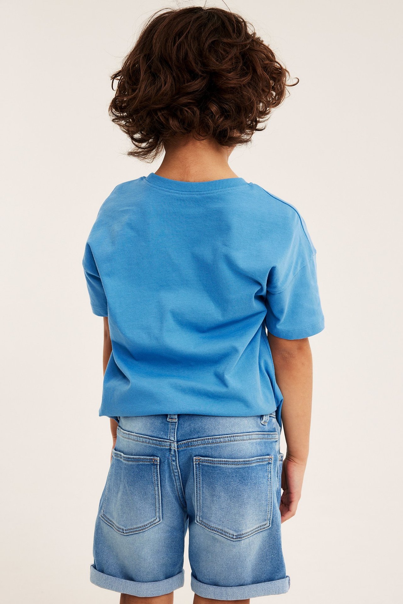 Lapsi takaapäin, yllään vaaleat denim-jogger shortsit käärityillä lahkeilla ja yksivärinen sininen t-paita.