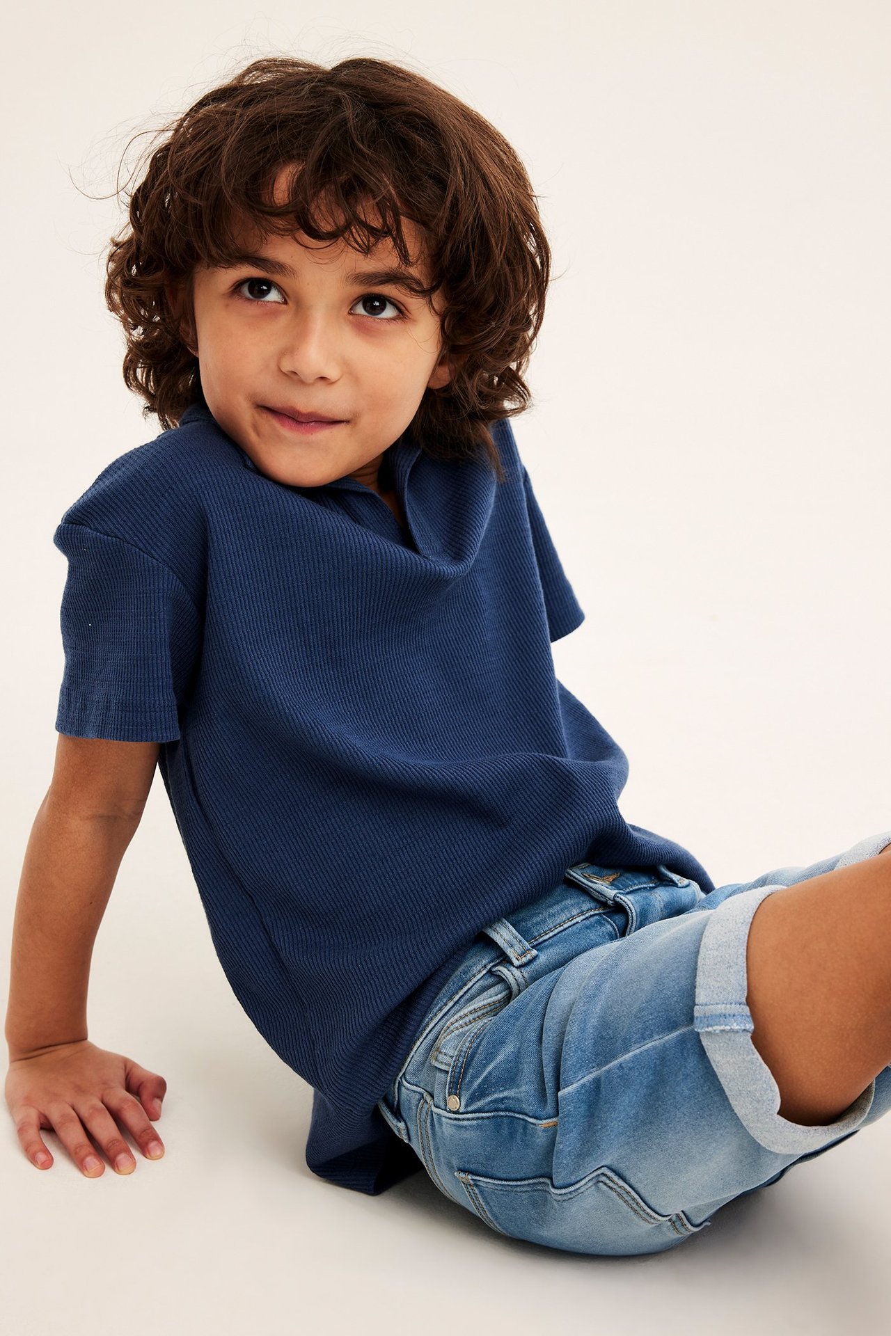 Edestä kuvattu lapsi yllään vaaleansiniset denim-trikooshortsit käärityillä lahkeilla ja tummansininen ribattu pikeepaita.