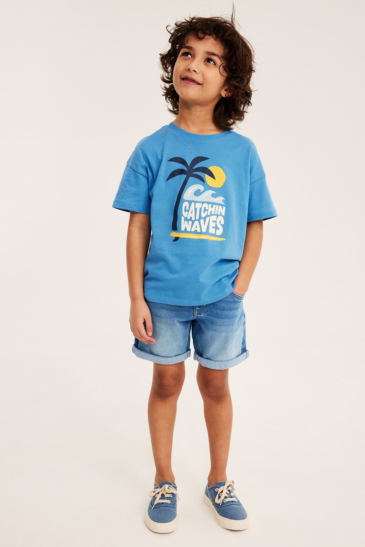 Helbild på ett barn i ljusa denimtrikåshorts, en blå mönstrad t-shirt och blå canvasskor.