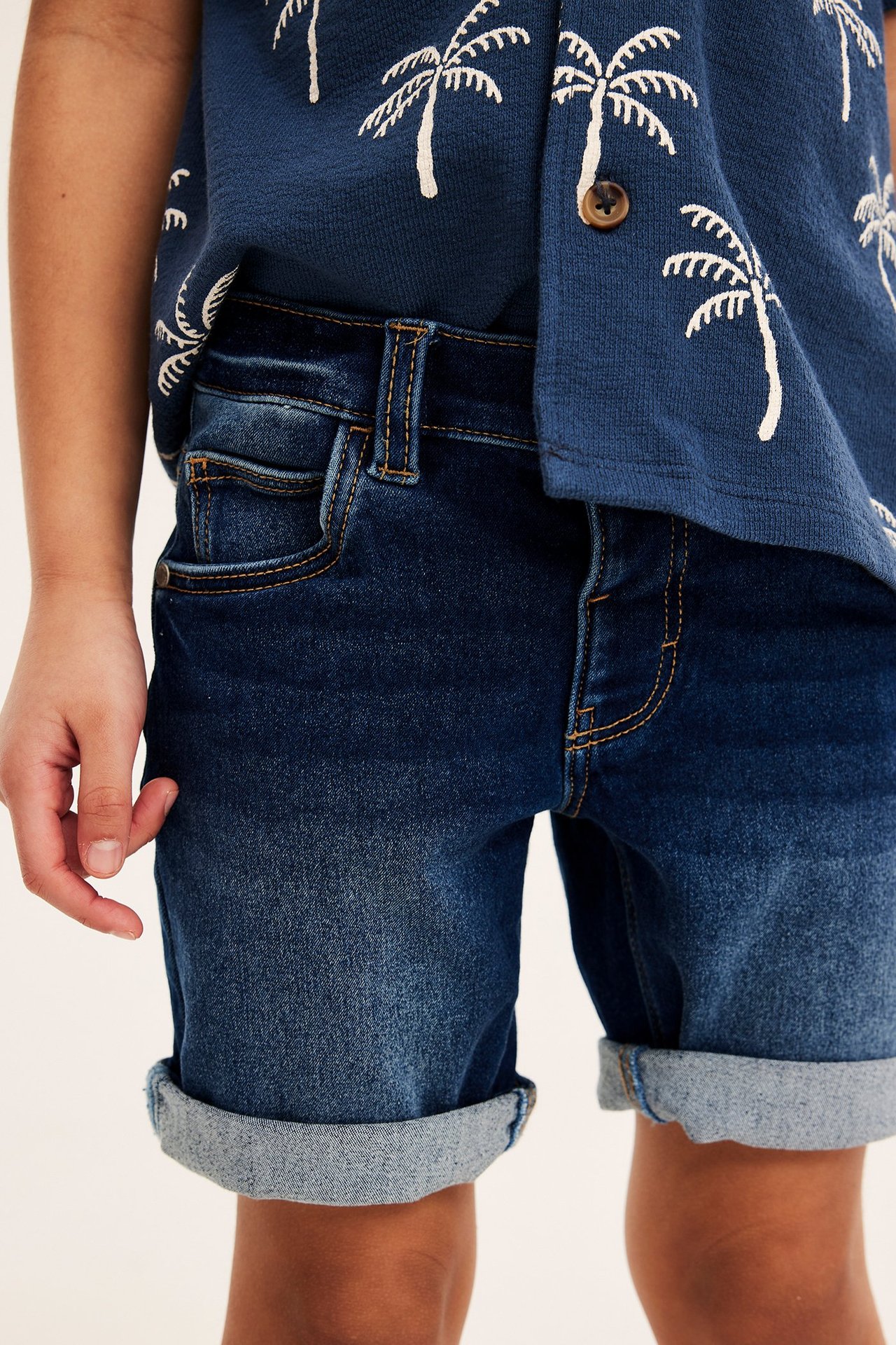 Frontvy av barn i mörkblå denimtrikåshorts med upprullade benslut och en mörkblå skjorta med palmträdsmönster.
