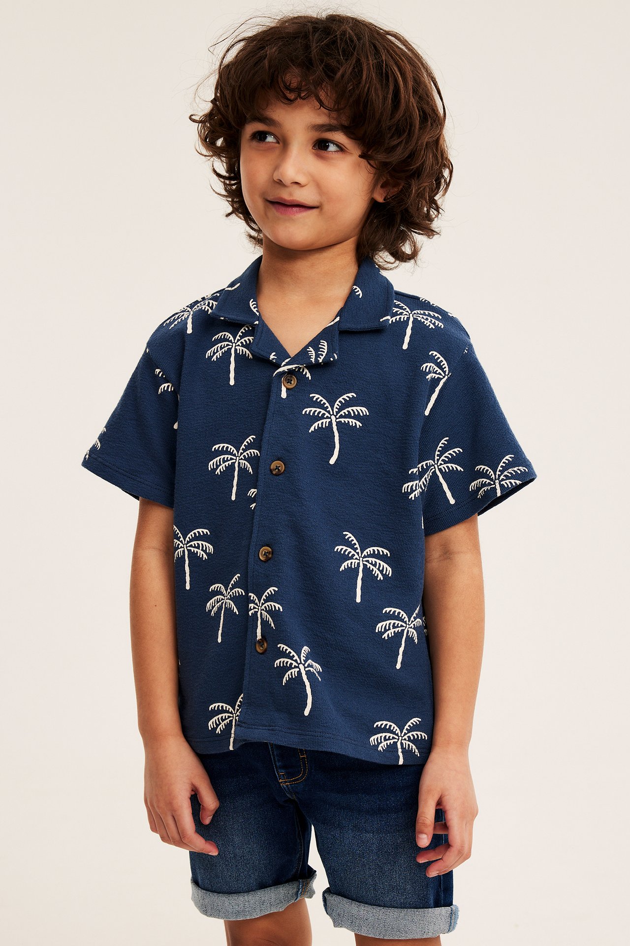 Frontvy: Ett barn bär mörkblå jeansshorts med uppvikta benslut och en mörkblå palmskjorta.