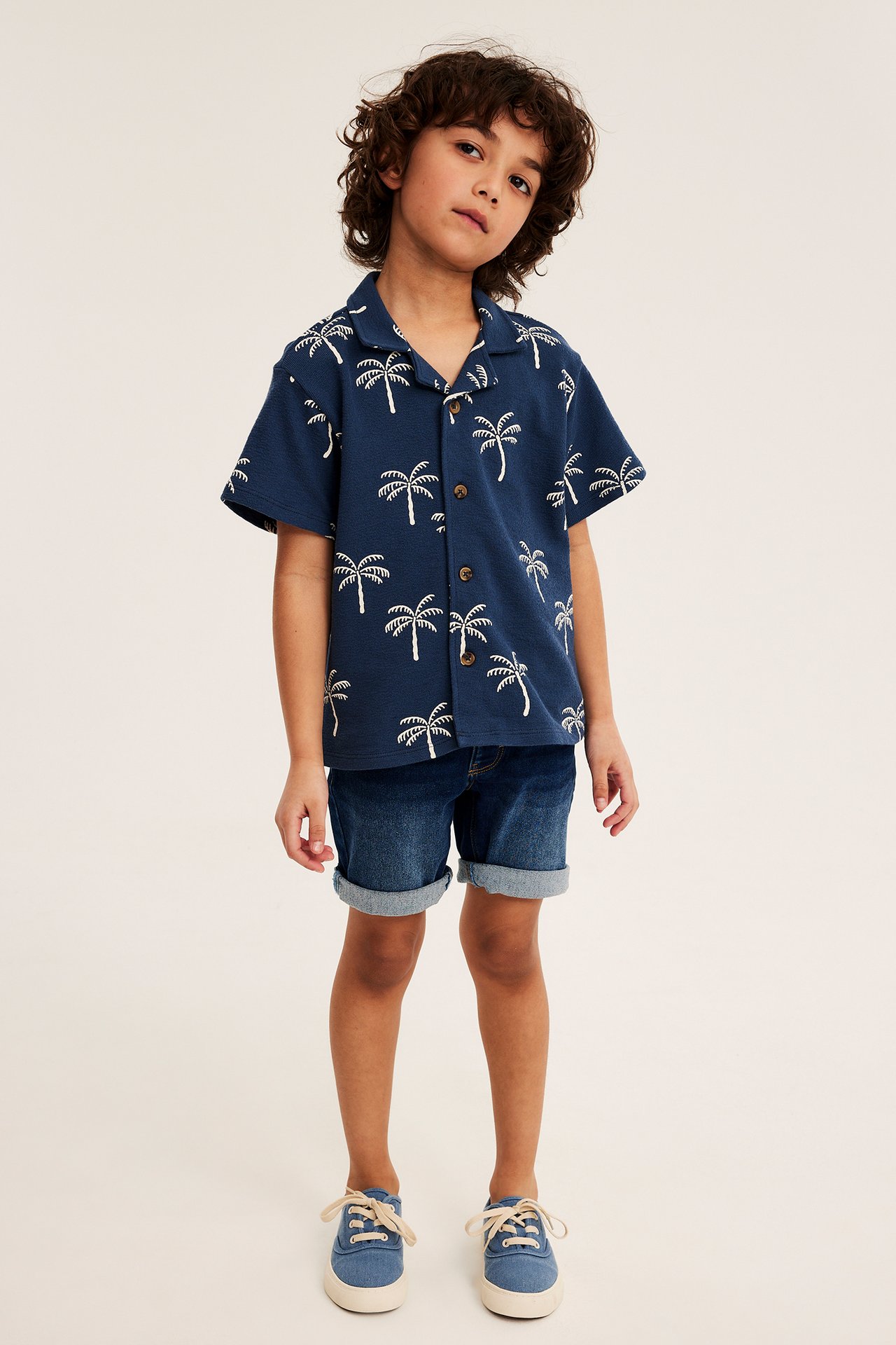 Framsida: Ett barn bär mörkblå denim jogger-shorts, en mörkblå skjorta med palmträdsmönster och blå sneakers.
