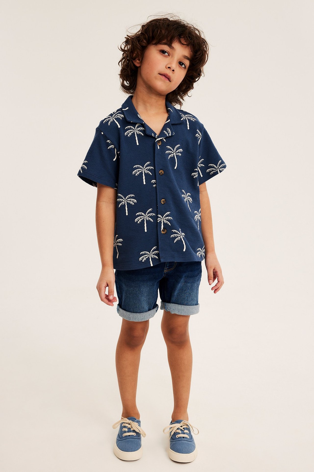 Widok z przodu dziecka: ciemnoniebieskie dżinsowe szorty jogger, niebieska koszula w palmy i niebieskie tenisówki.