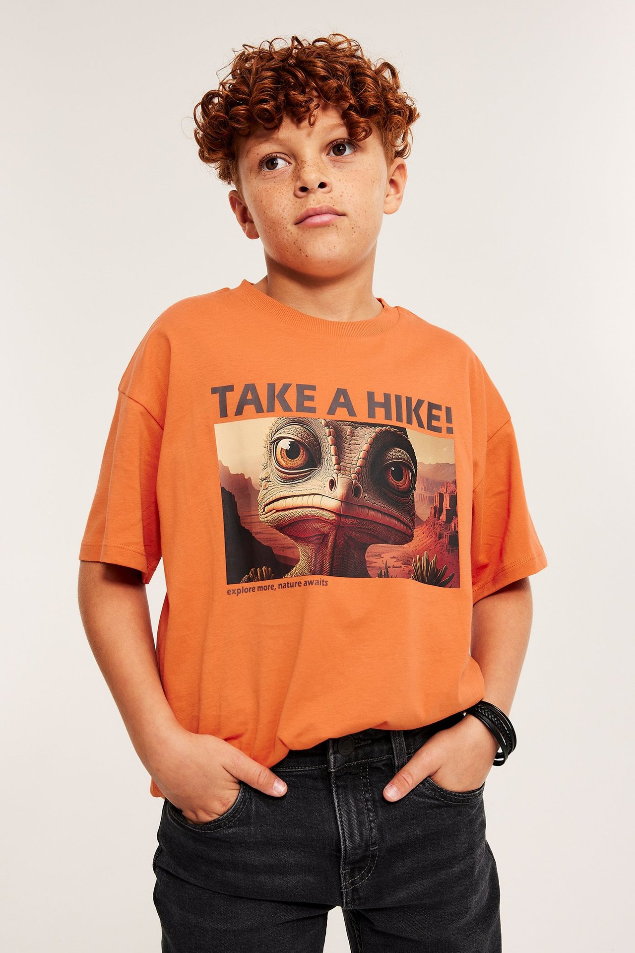 Barnhane i orange T-shirt med tryck, framifrån, över svart jeans.