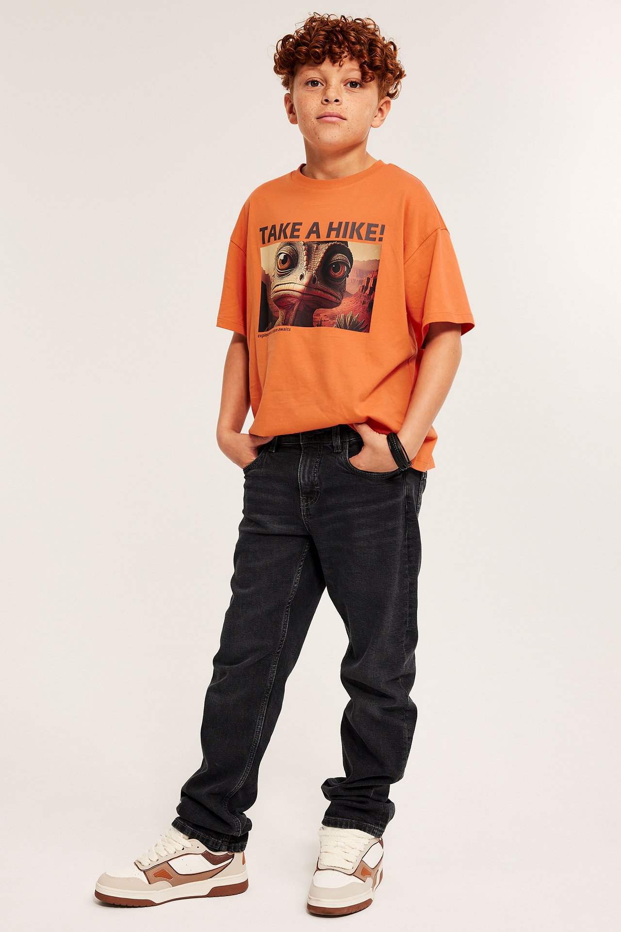 Chłopiec w pomarańczowym T-shircie z nadrukiem, czarne dżinsy, białe sneakersy, front.