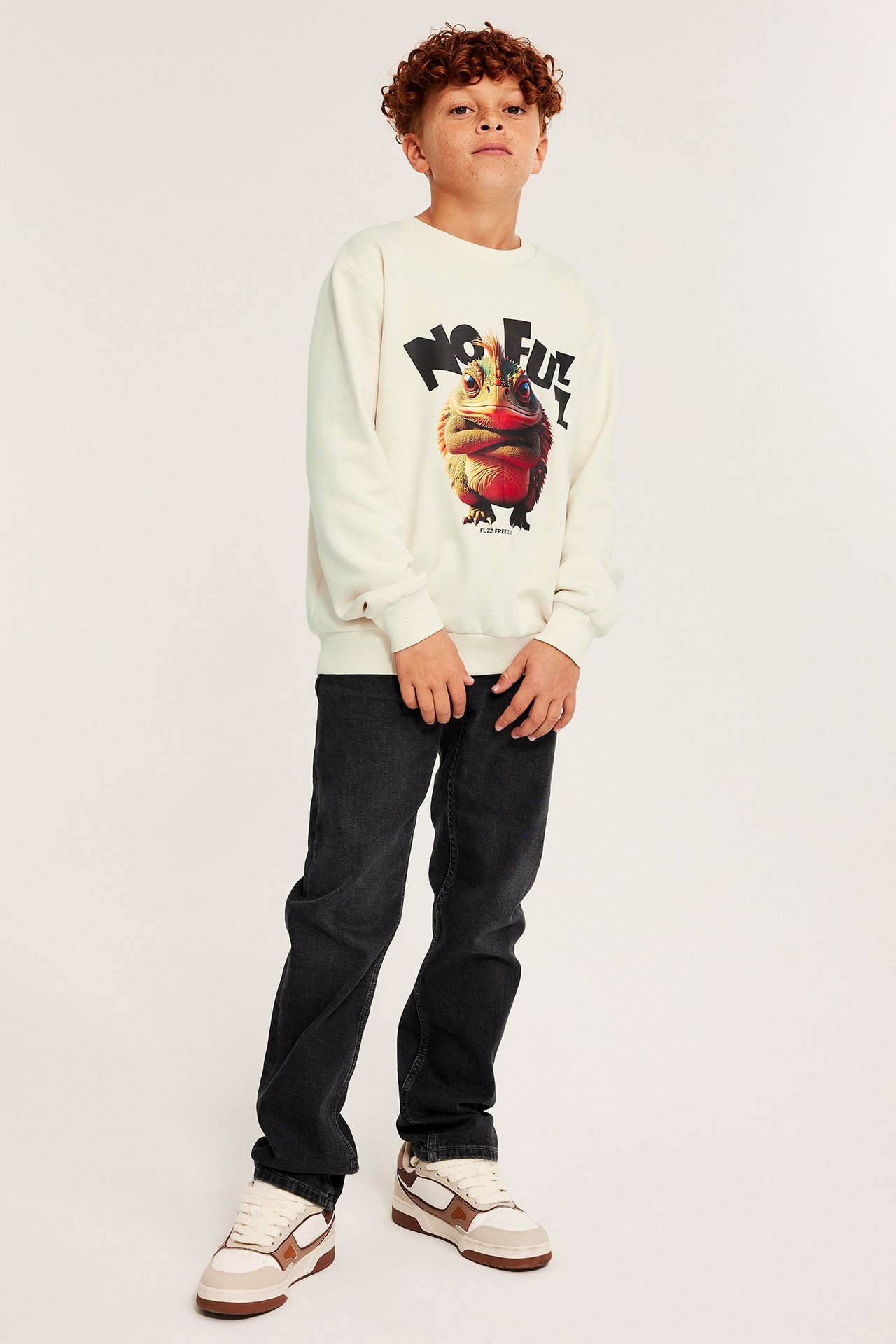 Dziecko w jasnobeżowej bluzie z dużym nadrukiem, ciemne dżinsy i białe sneakersy.