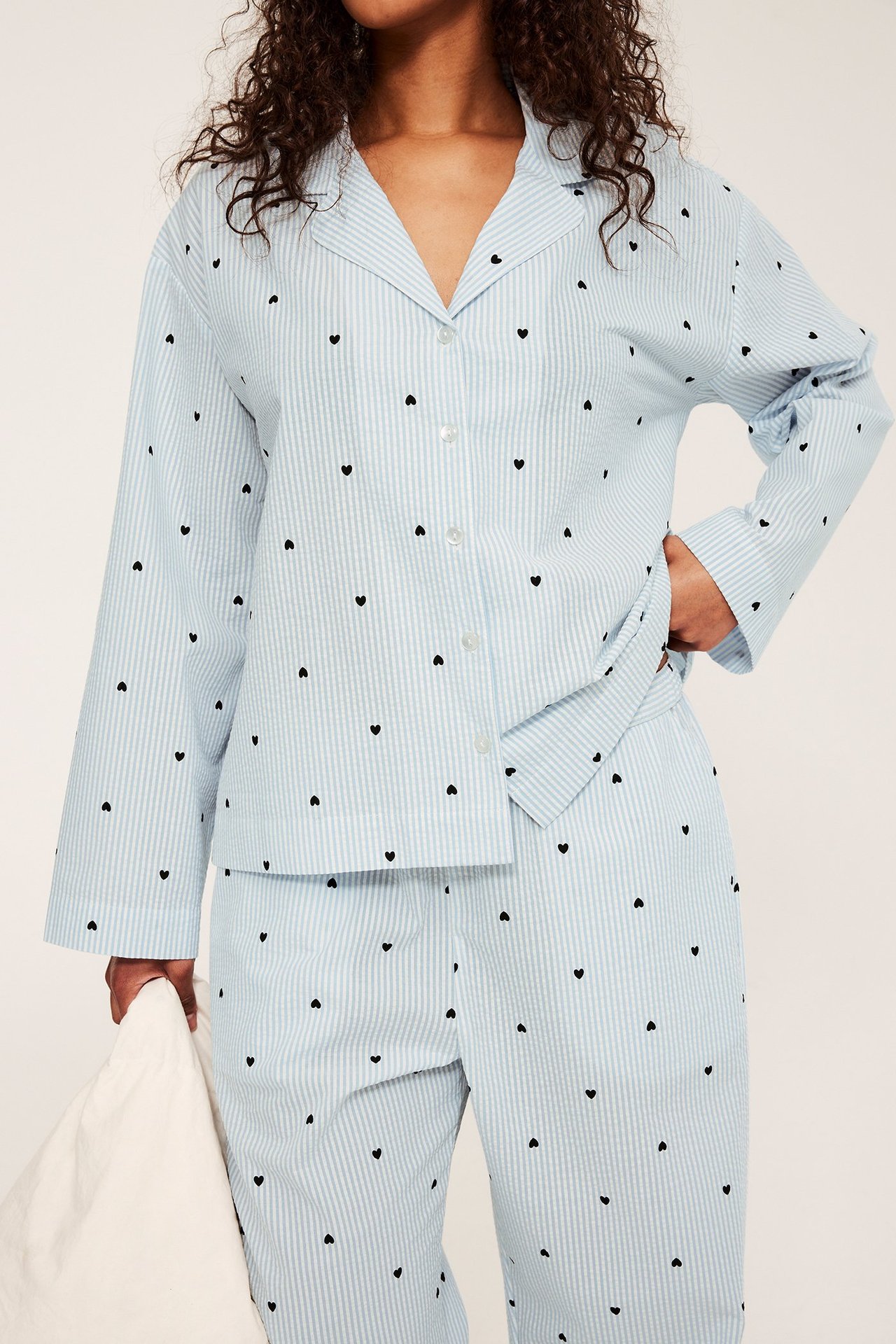 Edestä: nainen yllään vaaleansininen raidallinen seersucker-pyjama mustilla sydämillä, pitäen valkoista tyynyä.