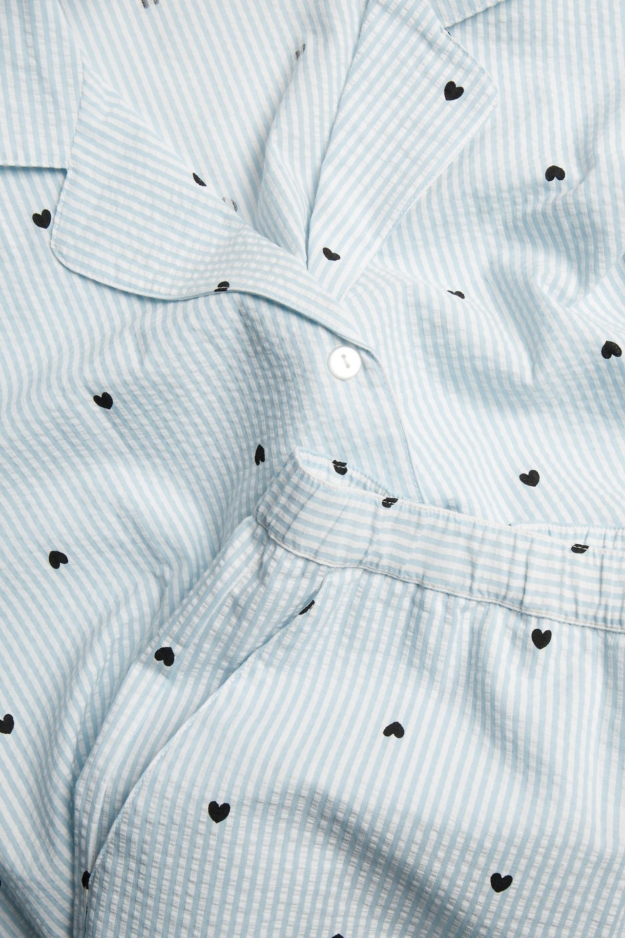 Zbliżenie jasnoniebieskiej, paskowanej piżamy seersucker z czarnym sercowatym wzorem i białym guzikiem.