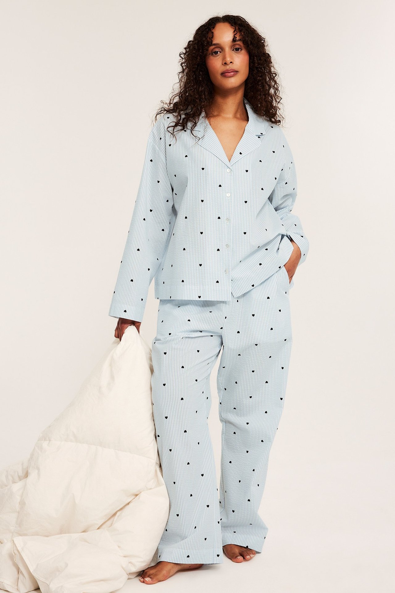 Edestäpäin, nainen yllään vaaleansininen raidallinen pyjamas mustilla sydämillä, pitäen valkoista peittoa.