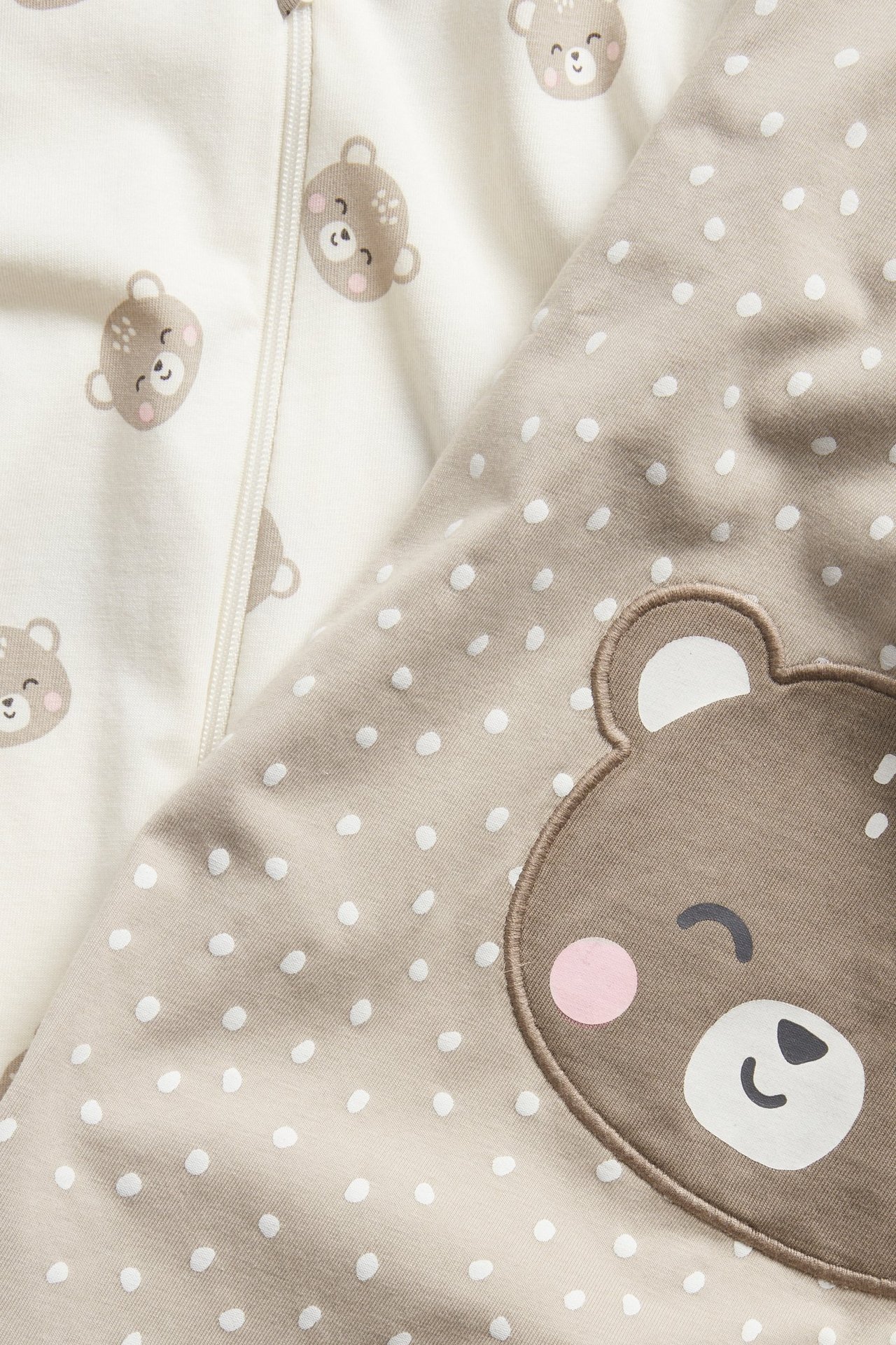 Nærbilde: To babyrompers. En beige med prikker, bjørneapplikasjon. En offwhite med bjørnetrykk, glidelås.