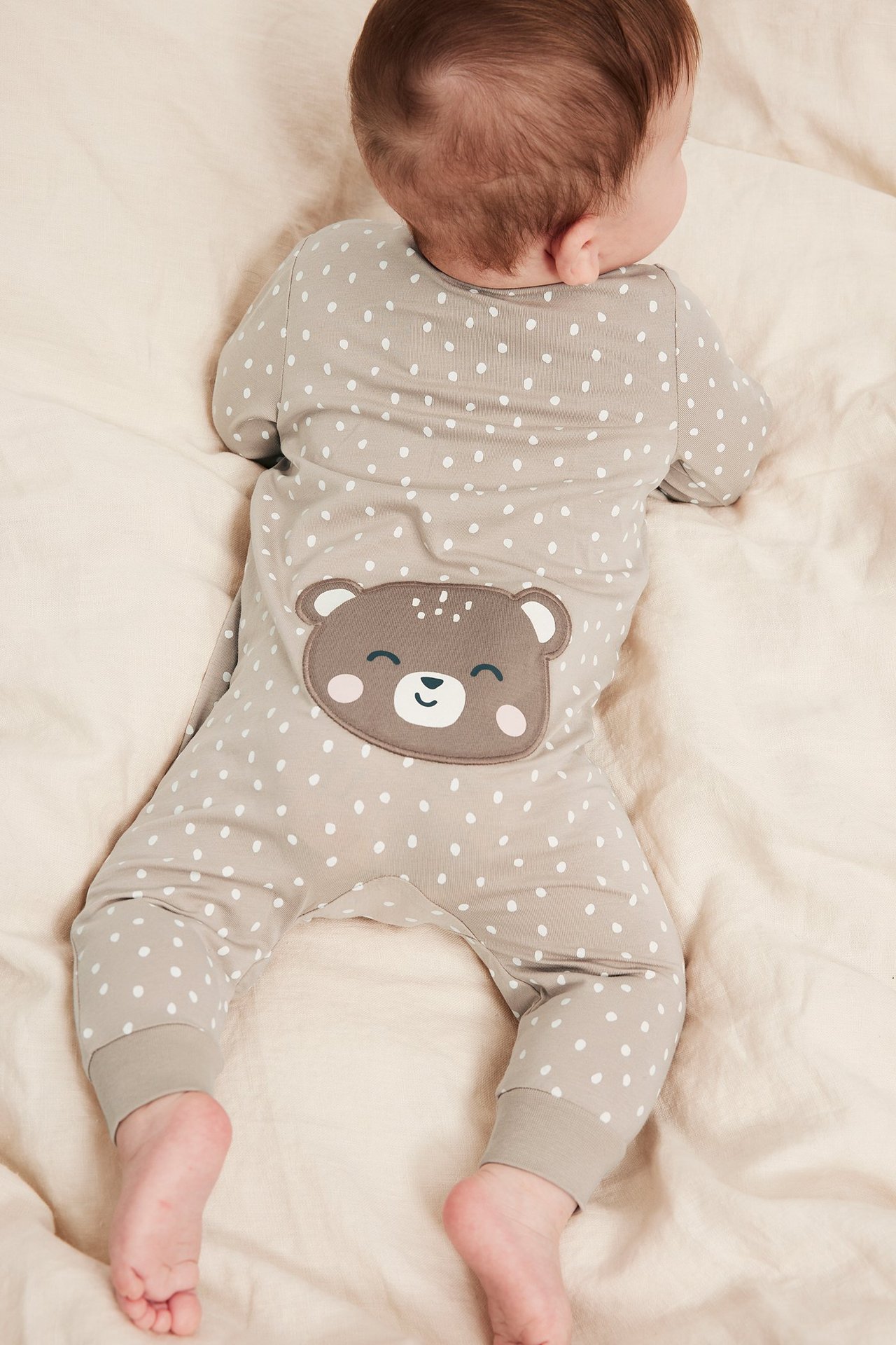 Widok z tyłu dziecka w jasnobeżowym pajacyku w kropki z brązową aplikacją misia na plecach.