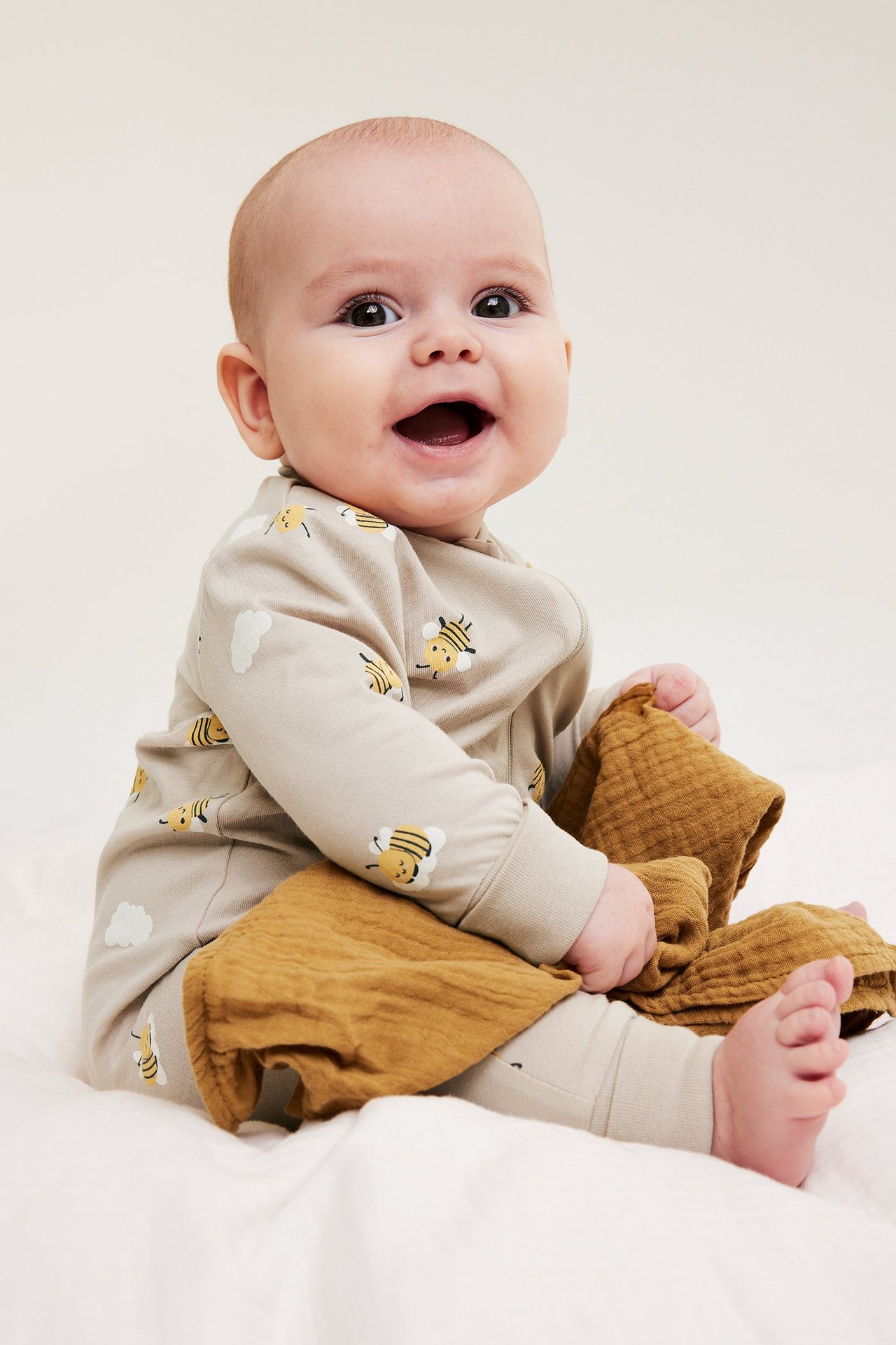 Uśmiechnięte dziecko w beżowym pajacyku w pszczoły i chmury, trzyma brązową pieluchę muślinową.