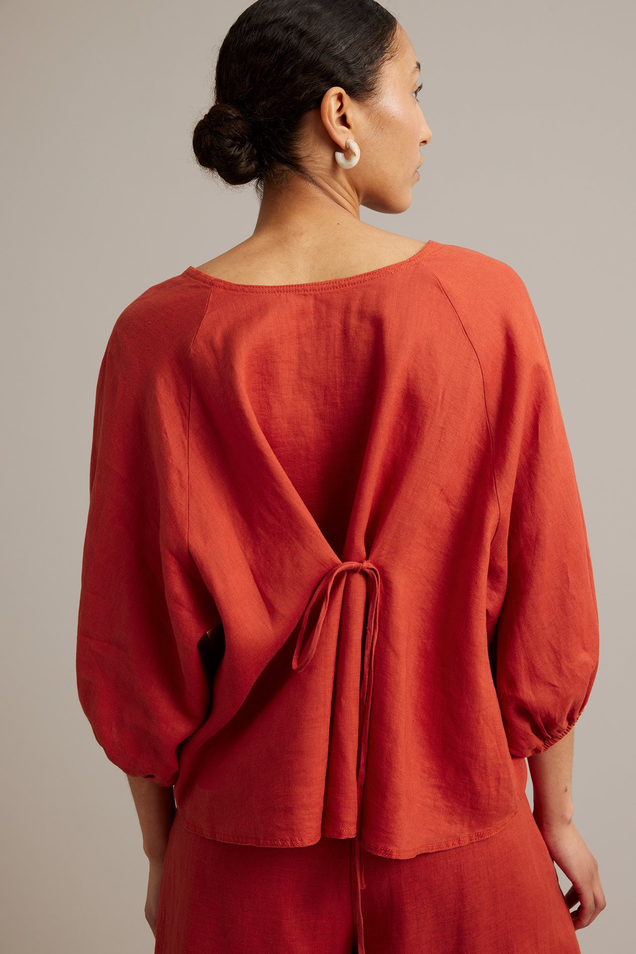 Ryggvy: kvinna i röd linneblus med vida raglanärmar, knytdetalj baktill och vita ringörhängen.