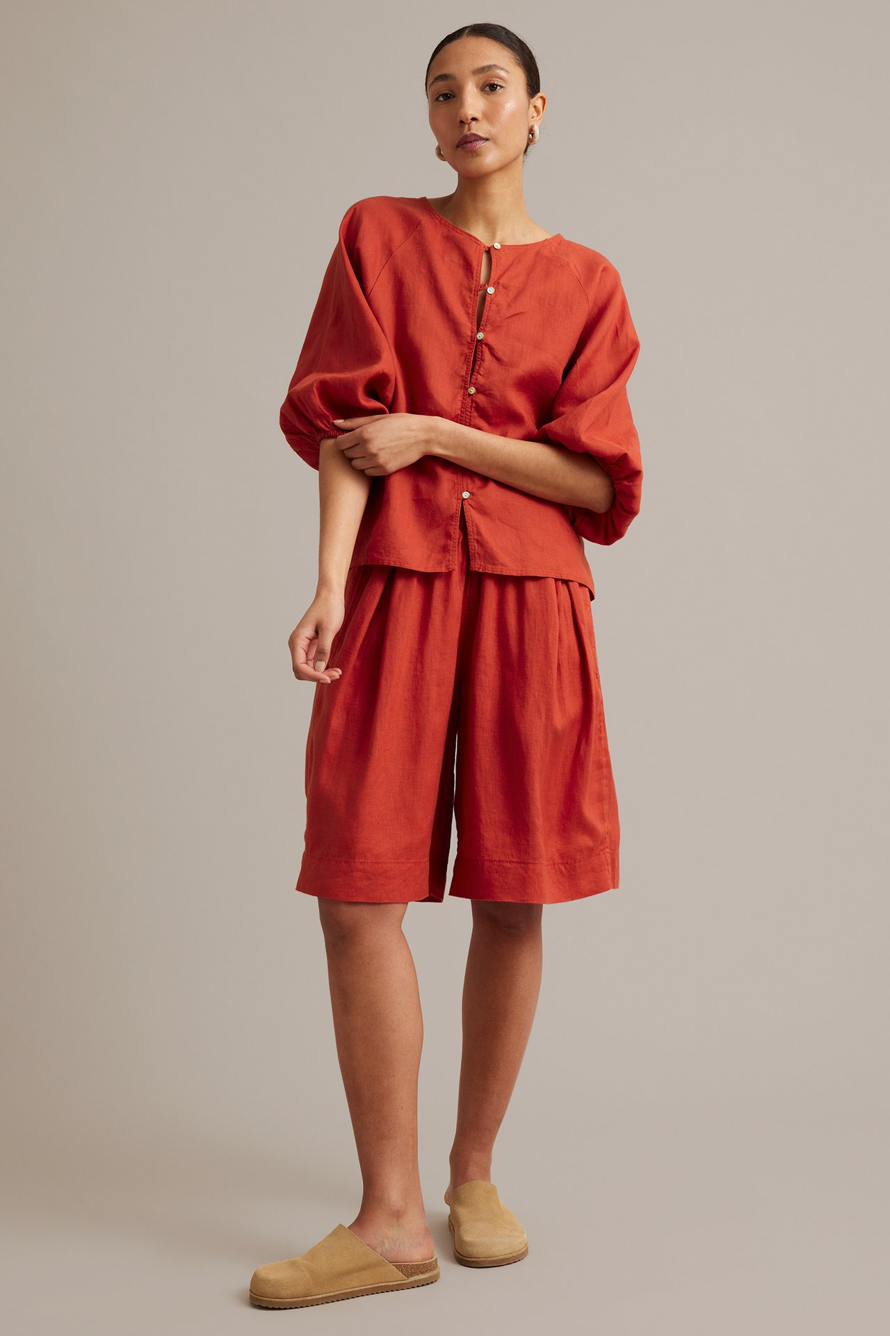 Forfra: kvinne i rød linbluse med raglanermer og knapper, matchende shorts og beige mulesko.