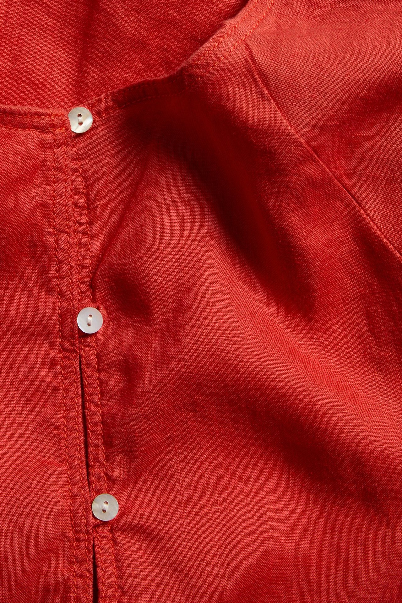 Zbliżenie czerwonej lnianej bluzki z okrągłym dekoltem i białą listwą guzikową.