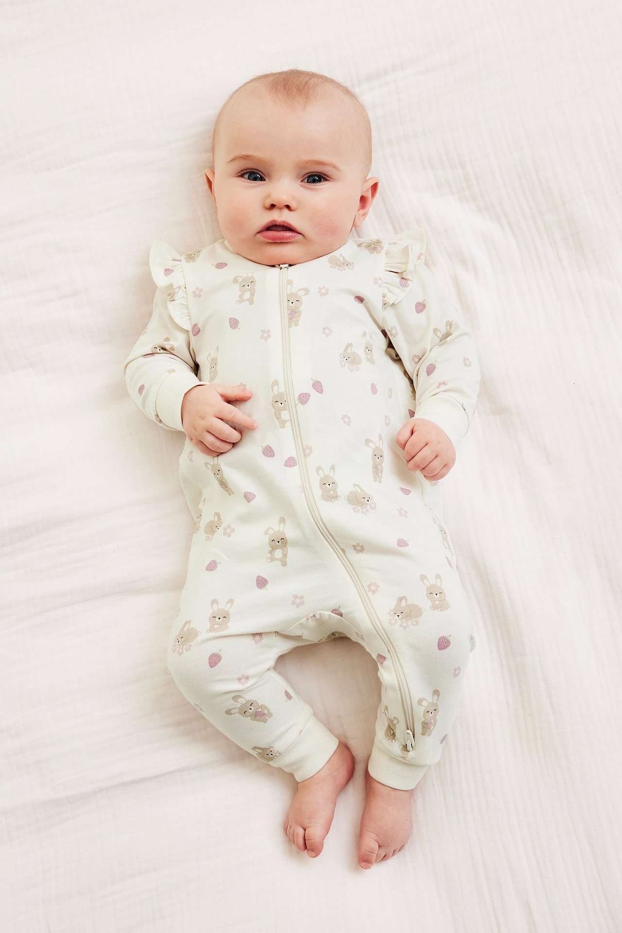 Z przodu: niemowlę w ecru pajacyku z królikami, truskawkami, falbankami na ramionach i dwukierunkowym zamkiem.