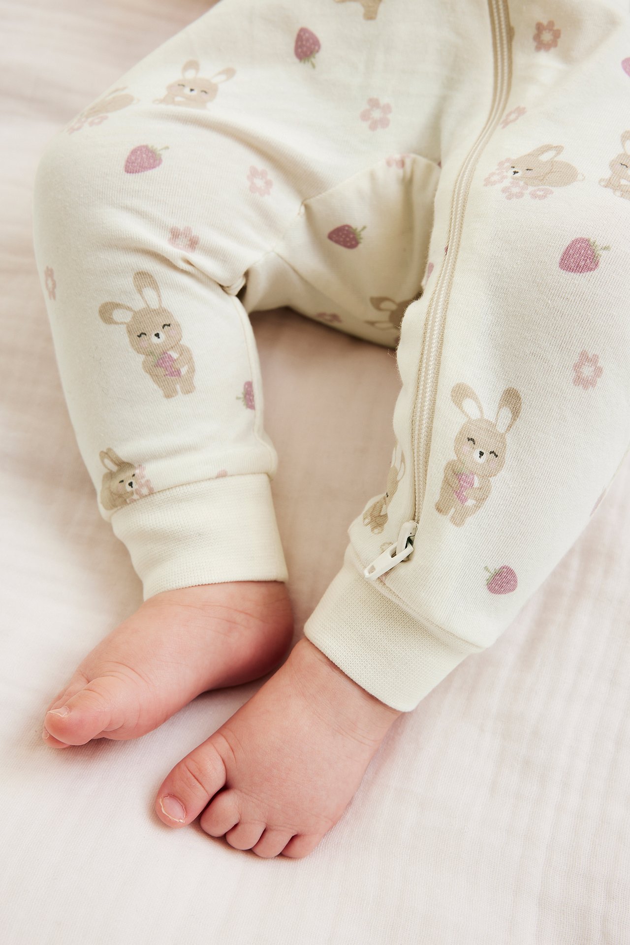 Nærbilde av babyben i offwhite heldress med kanin- og jordbærtrykk, med glidelås.
