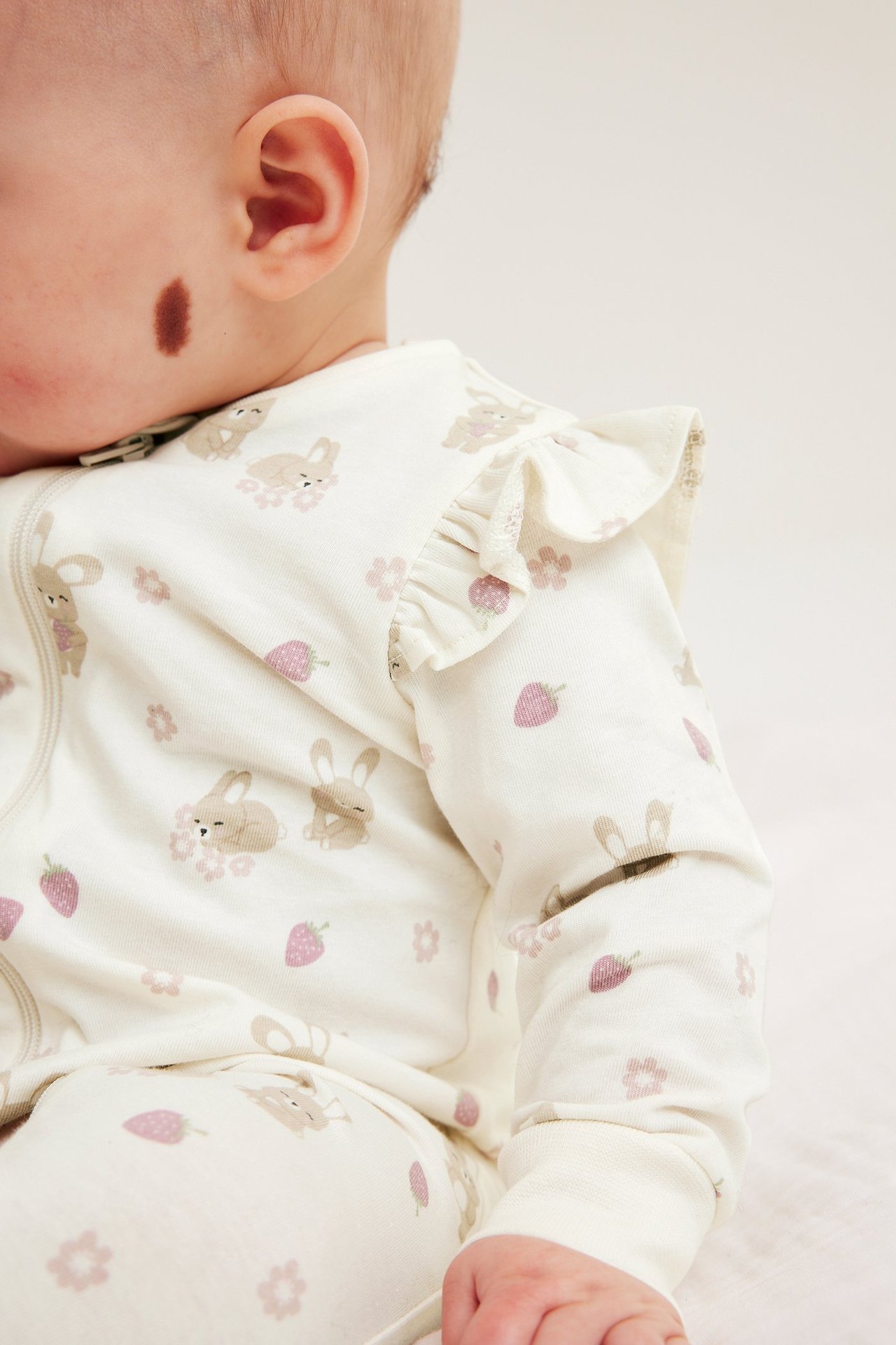 Boczny widok dziecka w białym wzorzystym pajacyku z króliczkami, truskawkami, falbankami i zamkiem.