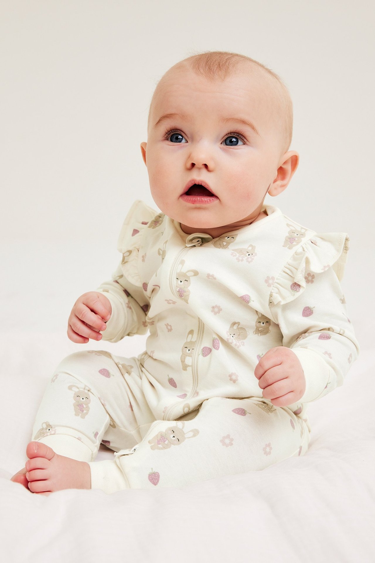 Widok z przodu: niemowlę w białym kombinezonie z wzorem królików, falbankami, dwukierunkowym zamkiem.