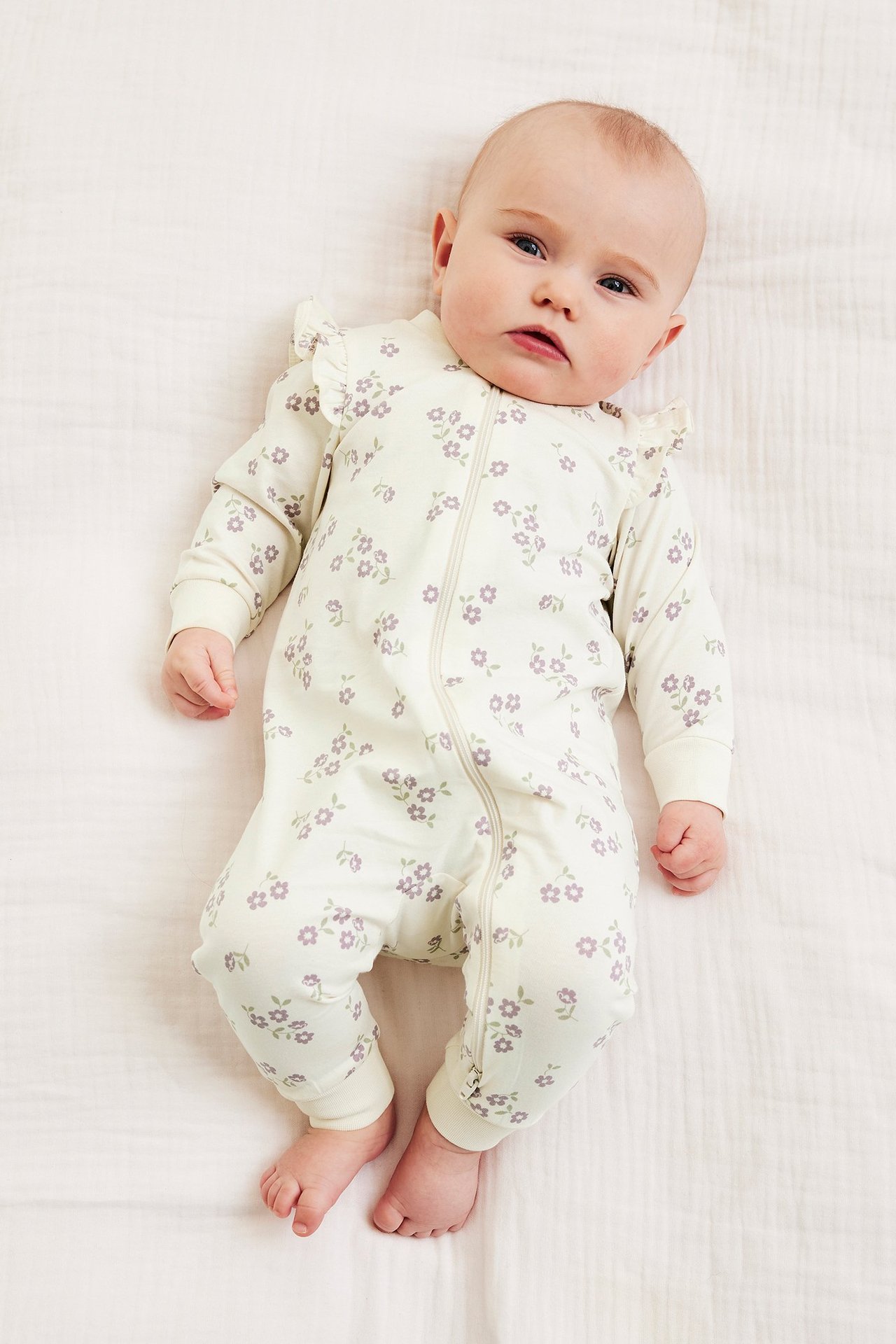 Crèmefärgad babyromper med lila blommönster, axelvolanger och tvåvägsdragkedja. Framifrån.