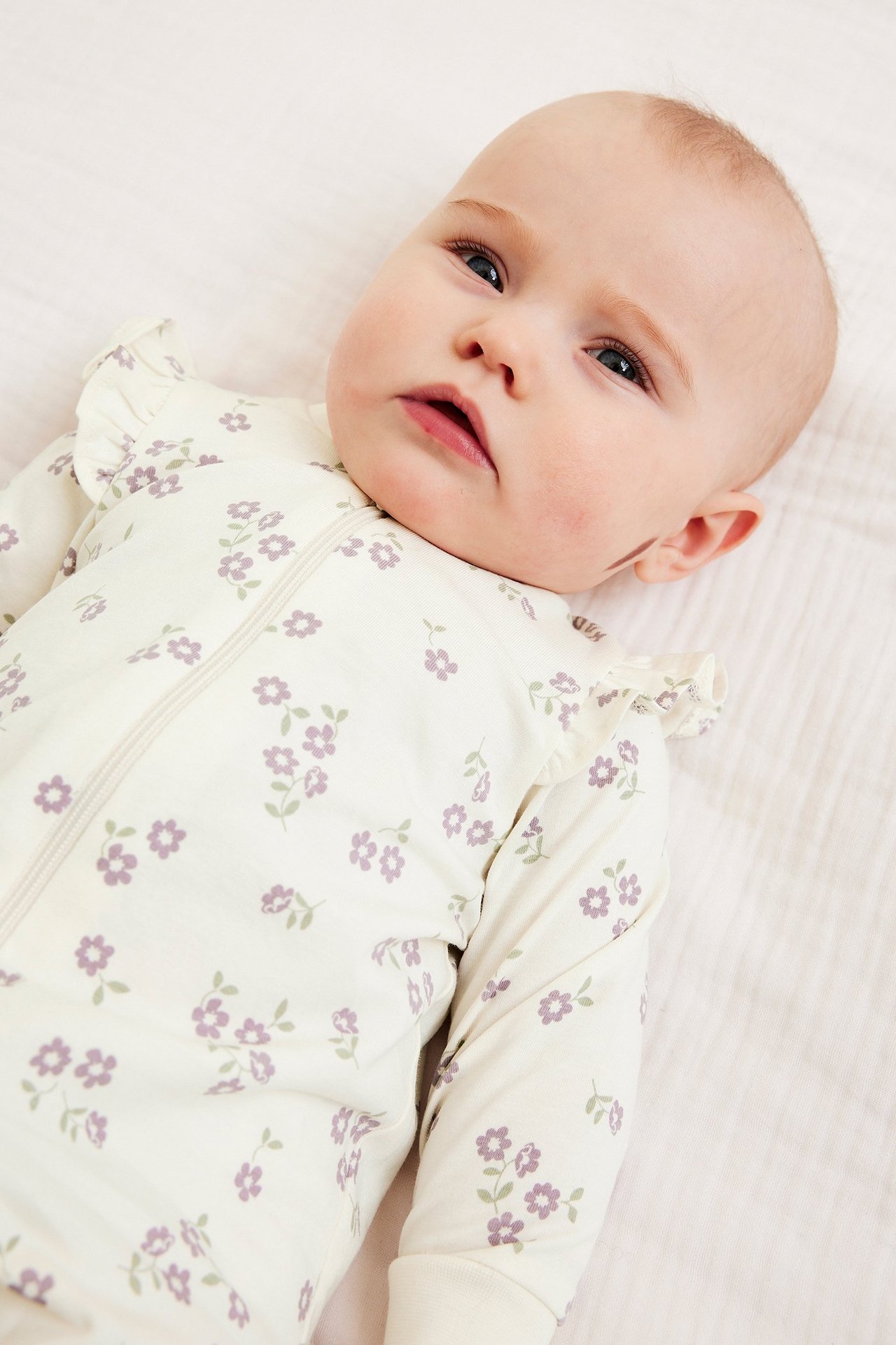 Zbliżenie: niemowlę w kremowym pajacyku z liliowym kwiatowym wzorem, dwukierunkowym zamkiem i falbankami na ramionach.