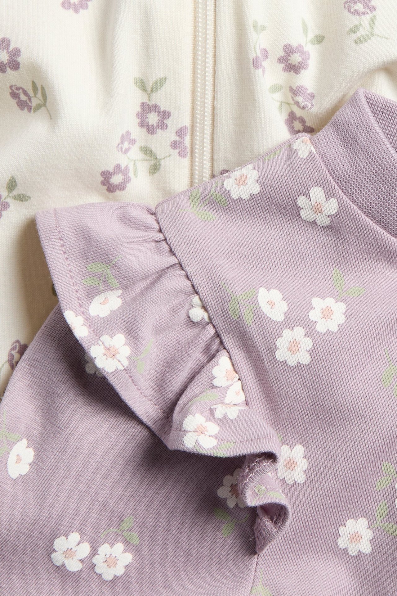 Zbliżenie dwóch pajacyków niemowlęcych: liliowego w kwiaty z falbankami i kremowego w kwiaty z zamkiem.