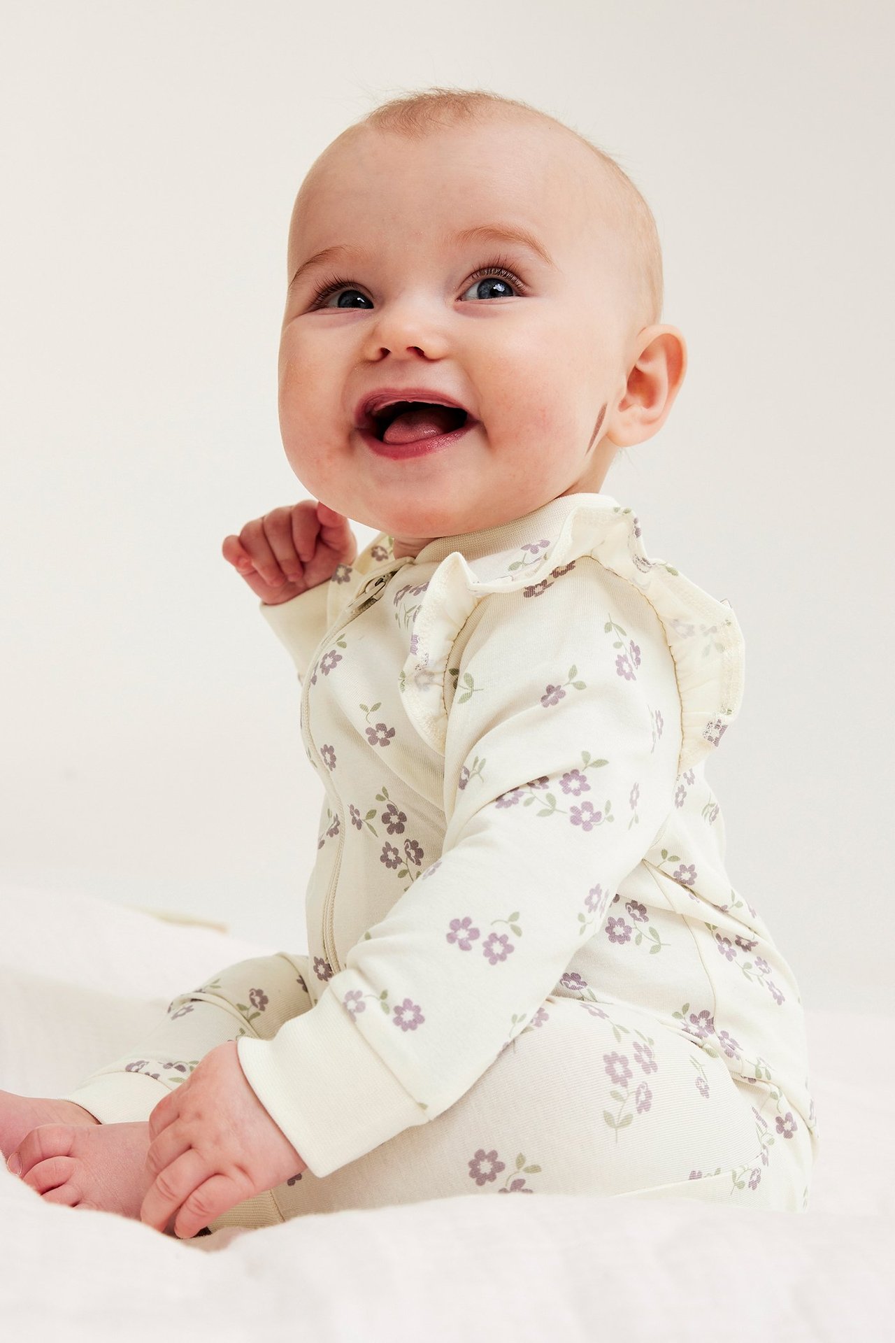 Widok z boku uśmiechniętego dziecka w kremowym pajacyku w fioletowe kwiaty z falbankami.