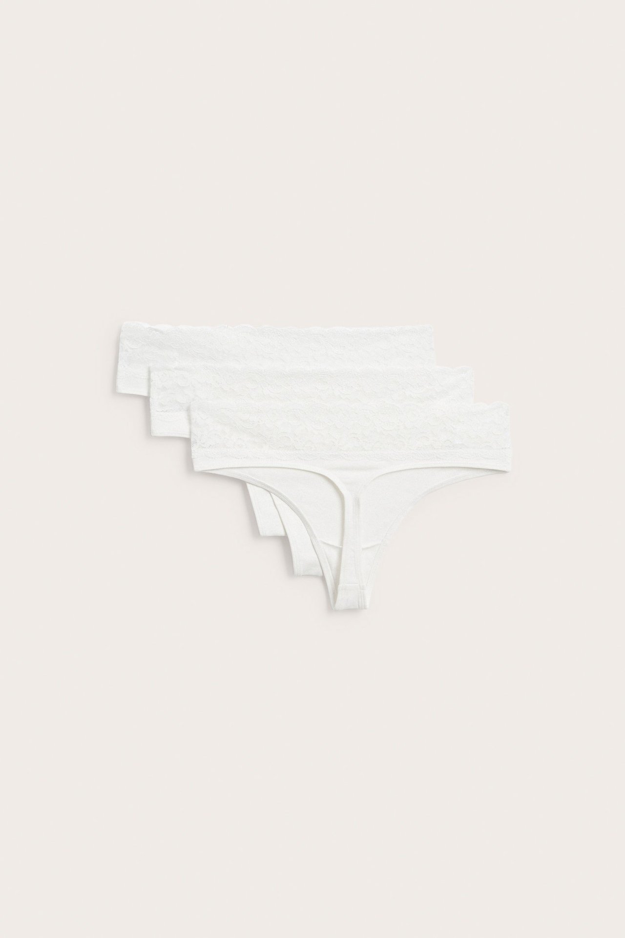 Flatt lay av tre hvite bomullsstringtruser med blonde i midjen, stablet.