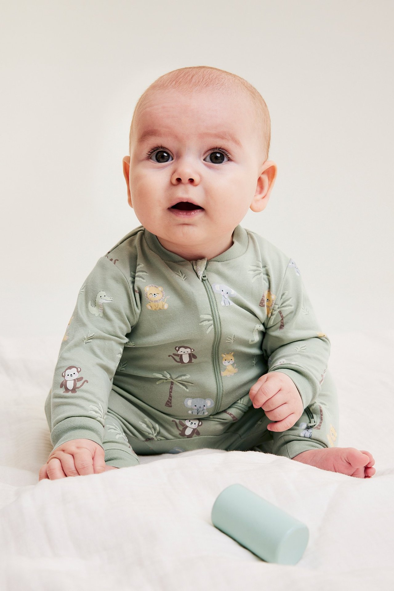 Edestä: Vauva vaaleanvihreässä eläinkuvioisessa haalarissa, kaksisuuntainen vetoketju, sinisen lelun luona.