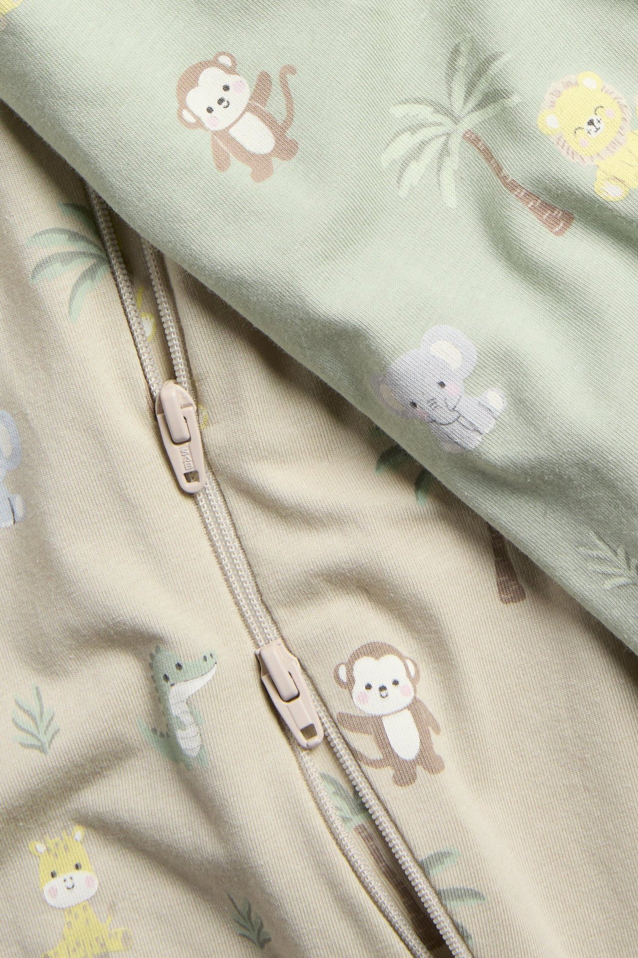 Zbliżenie dwóch pajacyków niemowlęcych, jasnozielonego i beżowego, z nadrukiem zwierzęcym i zamkiem dwukierunkowym.