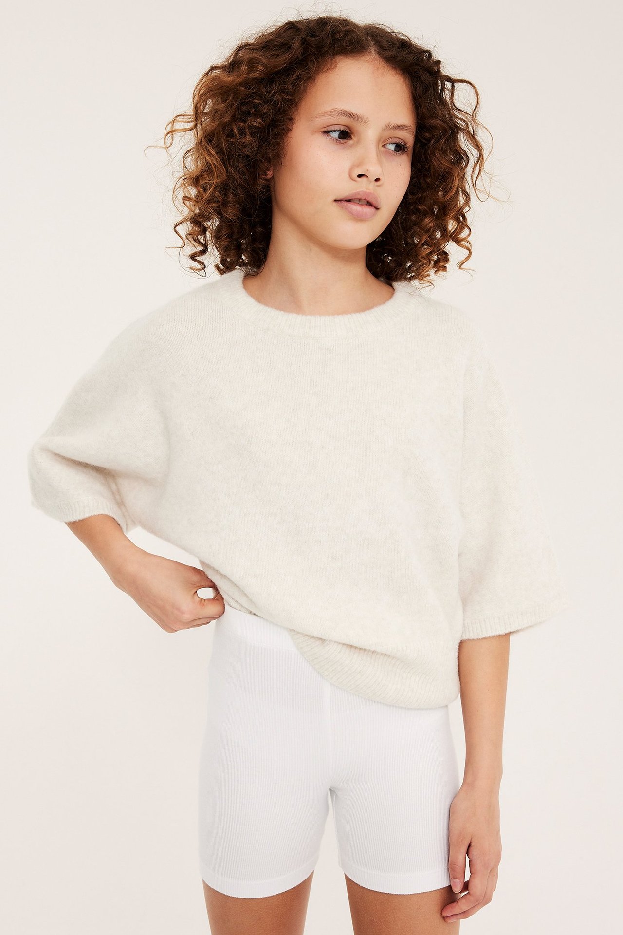 Lapsi edestä: valkoiset ribbineulotut pyöräilyshortsit ja löysä vaaleanbeige lyhythihainen pusero.