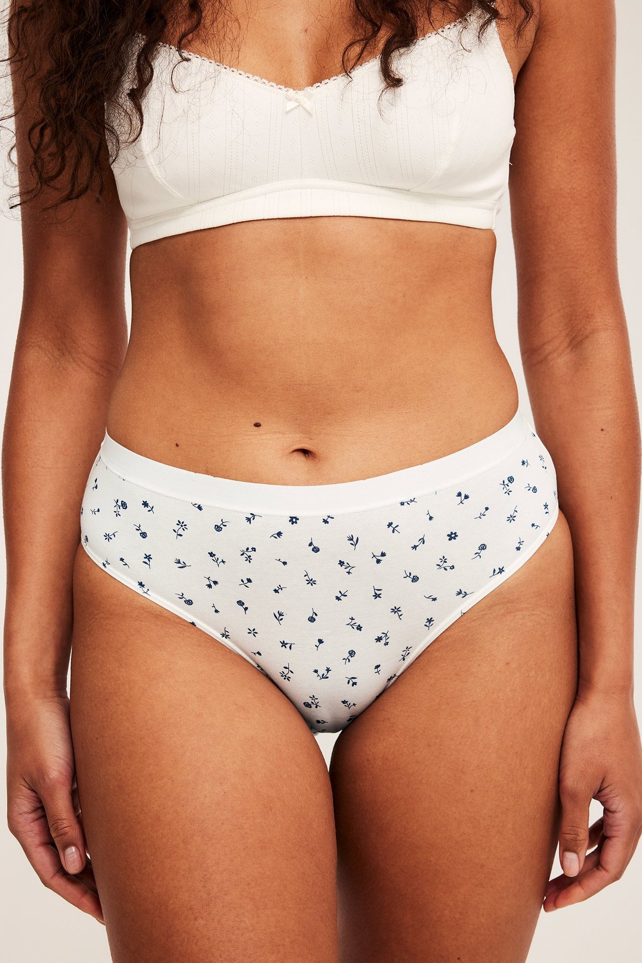 Edestä kuvattu nainen valkoisissa kukkakuvioisissa alushousuissa ja valkoisissa puuvillarintaliiveissä.