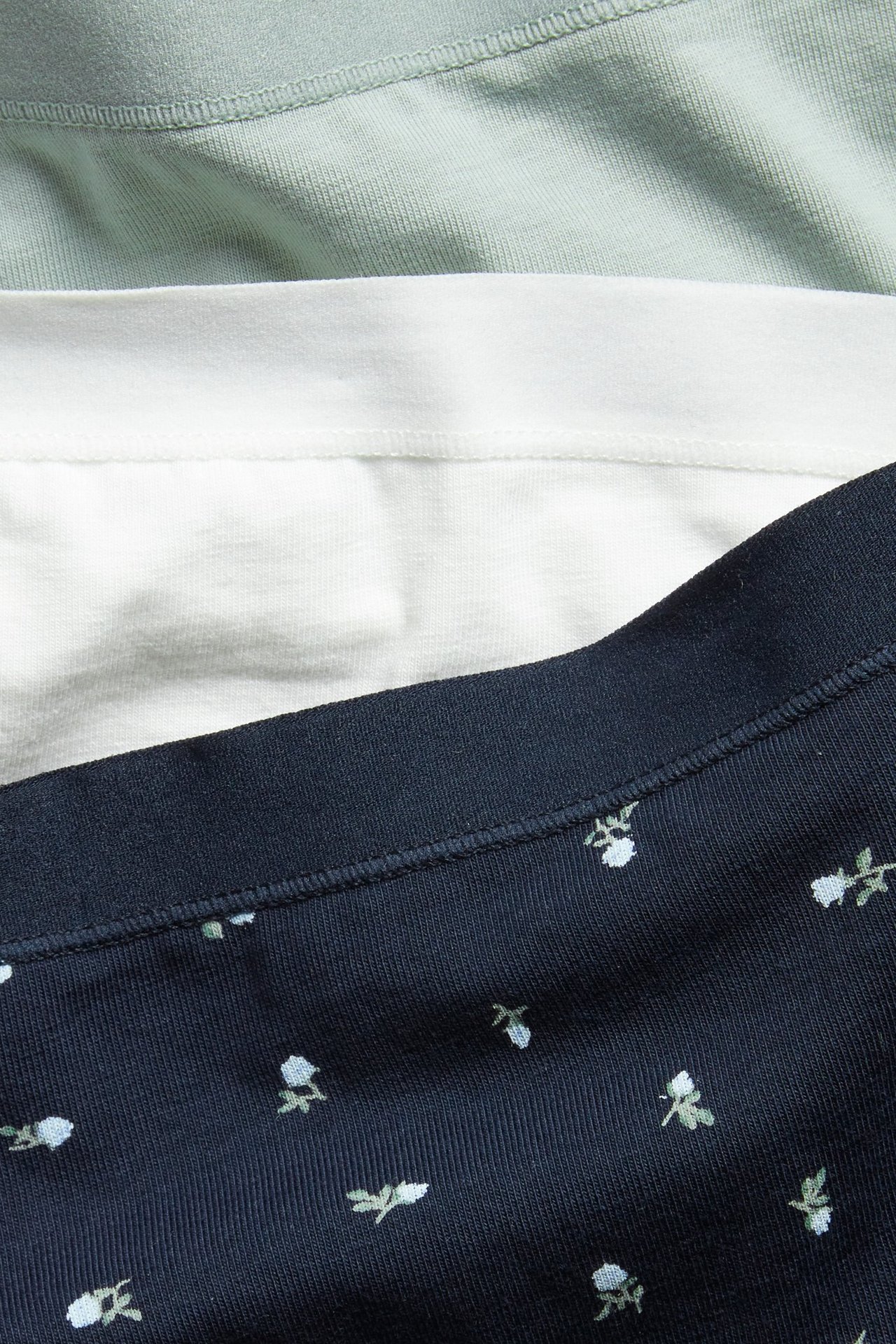 Nærbilde av en 3-pakning med bomullstruser for kvinner: en mørkeblå mønstret, en hvit og en lys grønn.