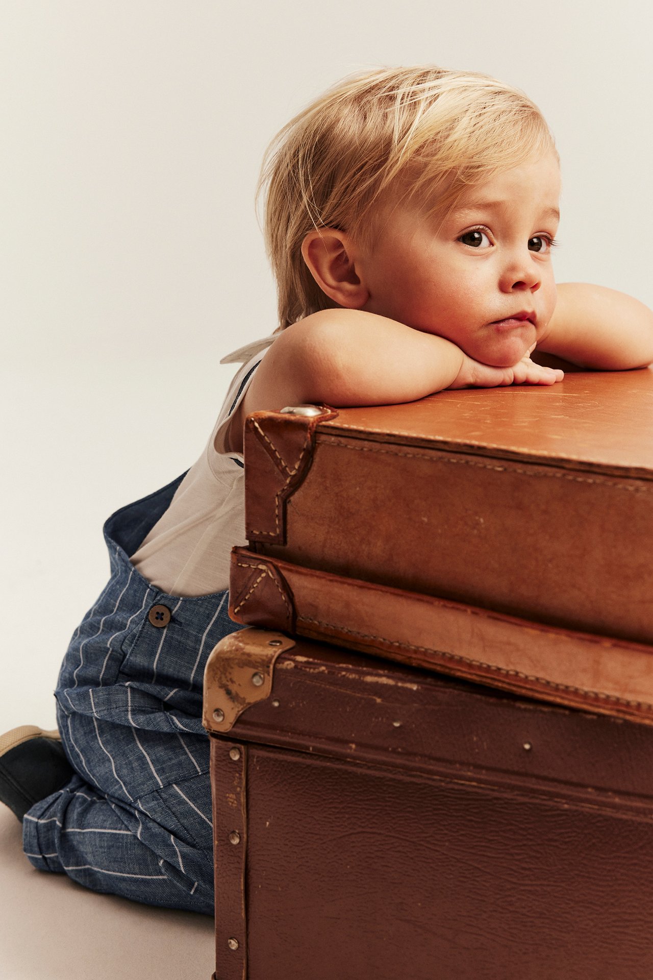 Lähikuva sivulta vauvasta siniraitaisissa puuvillahenkselihousuissa valkoisen topin päällä, nojaten matkalaukkuihin.