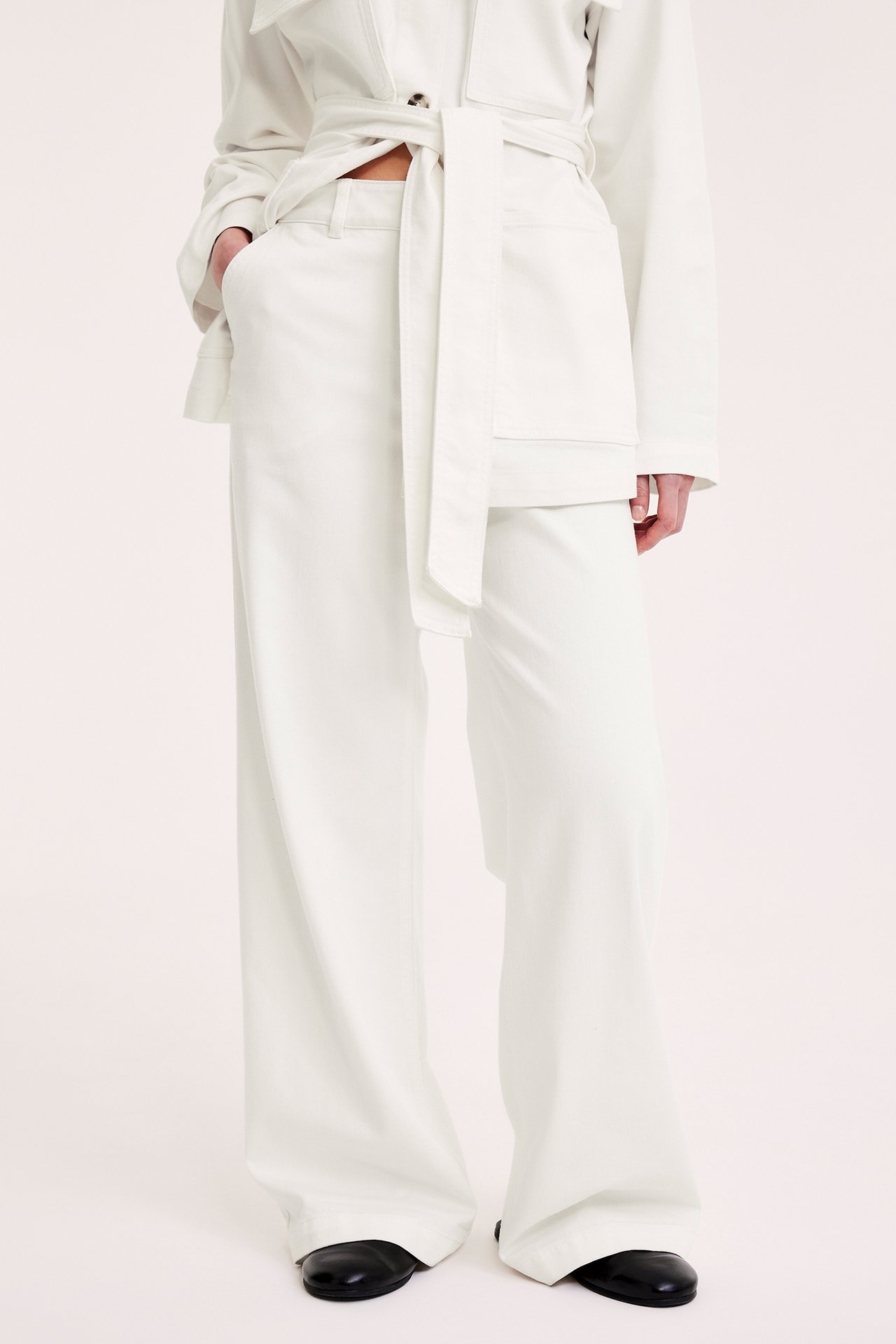 Edestä nainen yllään luonnonvalkoiset leveät tvillihousut, vyöllinen takki ja mustat kengät.