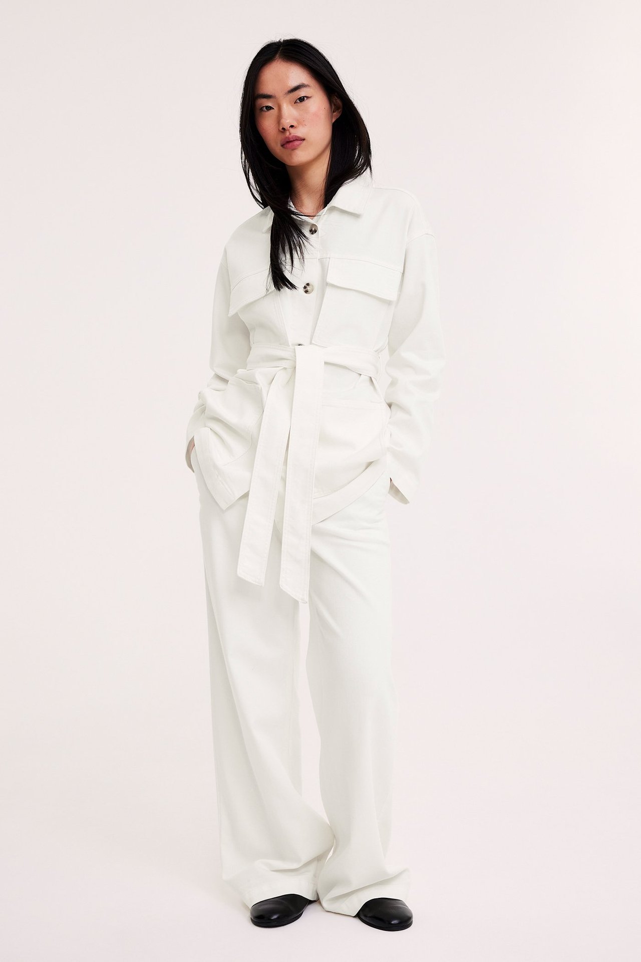 Nainen off-white leveälahkeisissa twill-housuissa, yhteensopivassa vyötärösolmitussa takissa ja mustissa matalissa kengissä.