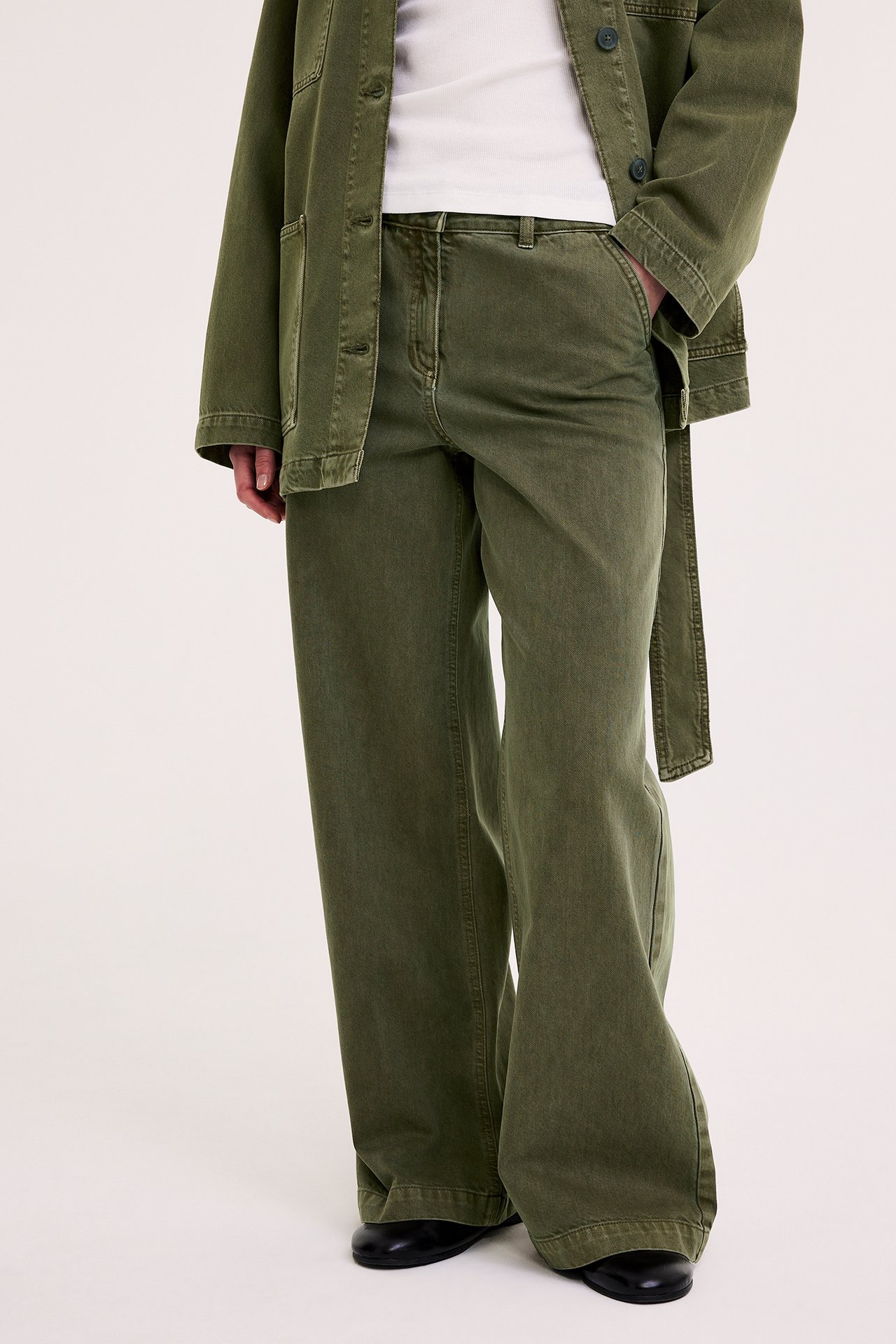 Edestä: nainen vihreissä leveälahkeisissa puuvillatvillihousuissa, valkoisessa t-paidassa, yhteensopivassa takissa, mustat kengät.