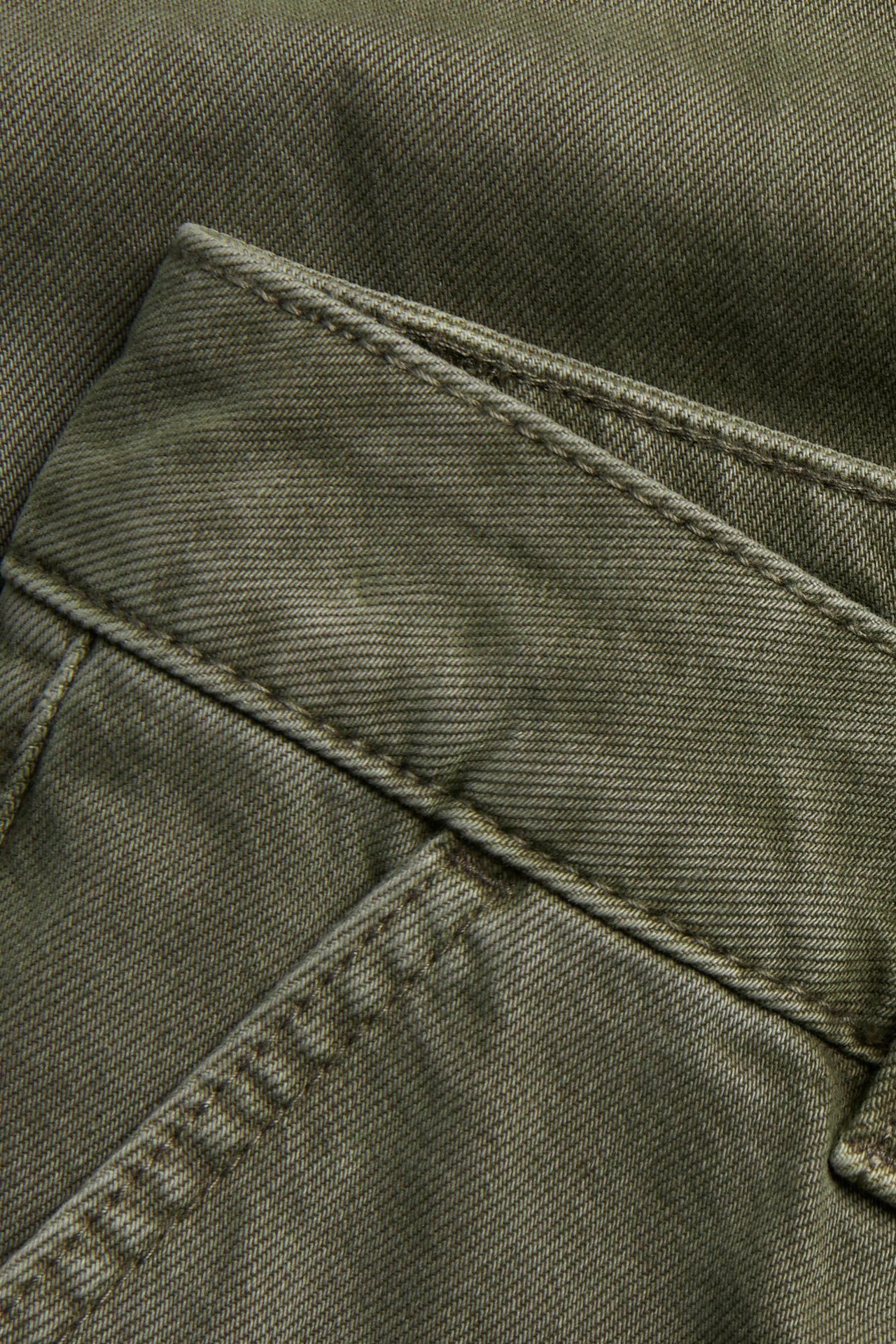 Zbliżenie zielonych twillowych spodni bawełnianych, widać teksturę, szwy i detal kieszeni.