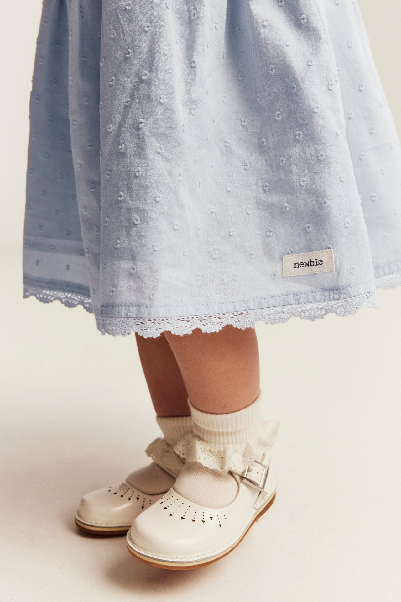 Närbild av ett barn i ljusblå prickig klänning med spetskant, vita volangstrumpor och vita spänneskor.