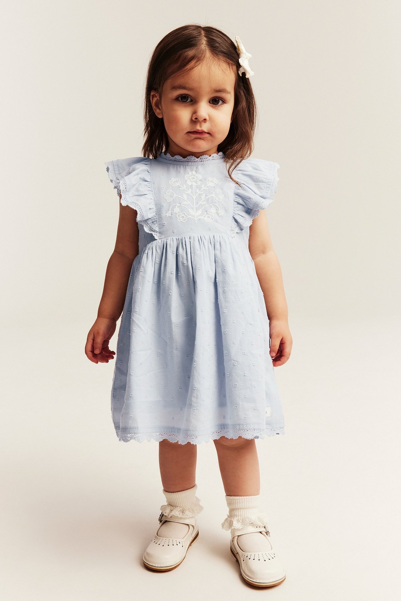 Edestä: Lapsi vaaleansinisessä brodeeratussa puuvillamekossa, jossa on röyhelöhihat, valkoiset rypytetyt sukat ja Mary Jane -kengät.