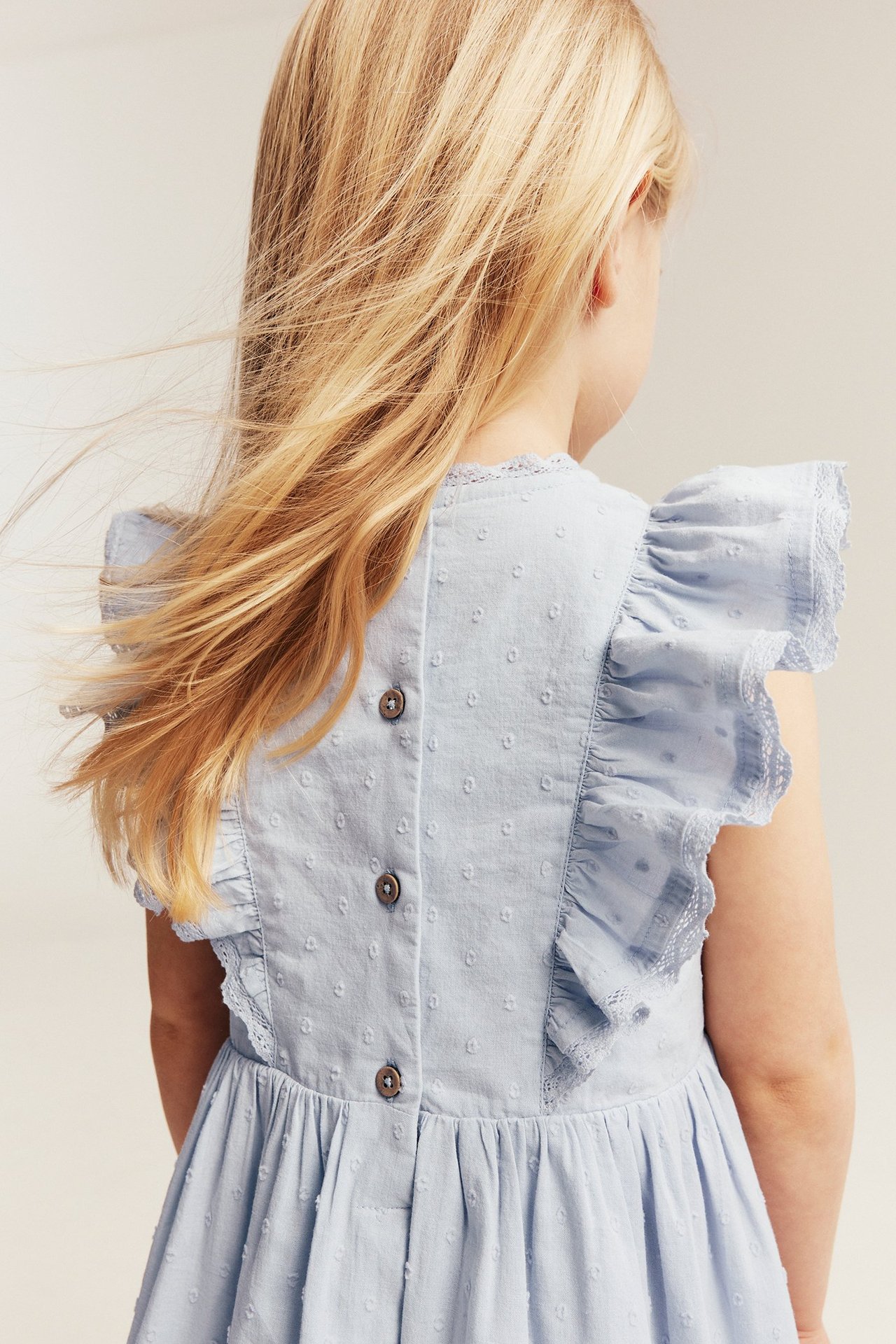 Bakfra: Barn i en lyseblå vevd bomullskjole med prikkstruktur, rysjeermer med blonder og knapper i ryggen.