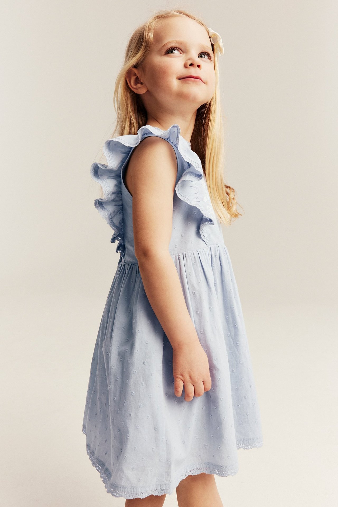 Sidevisning av et barn i lyseblå bomullskjole med prikkstruktur, volangermer og blondekant nederst.