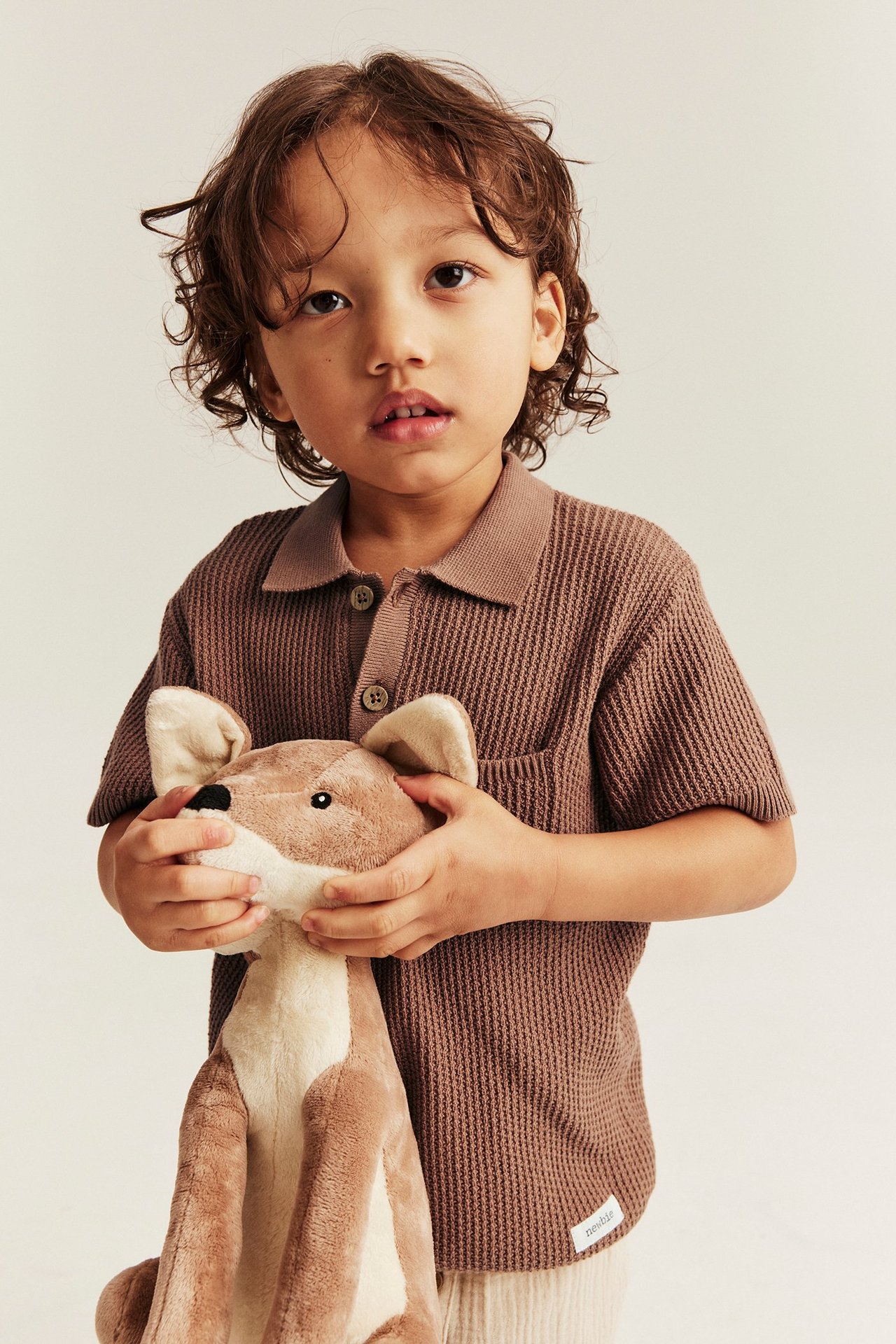 Frontvy: Ett barn i en brun stickad kortärmad pikétröja med ficka, håller i en mjukdjursleksak.