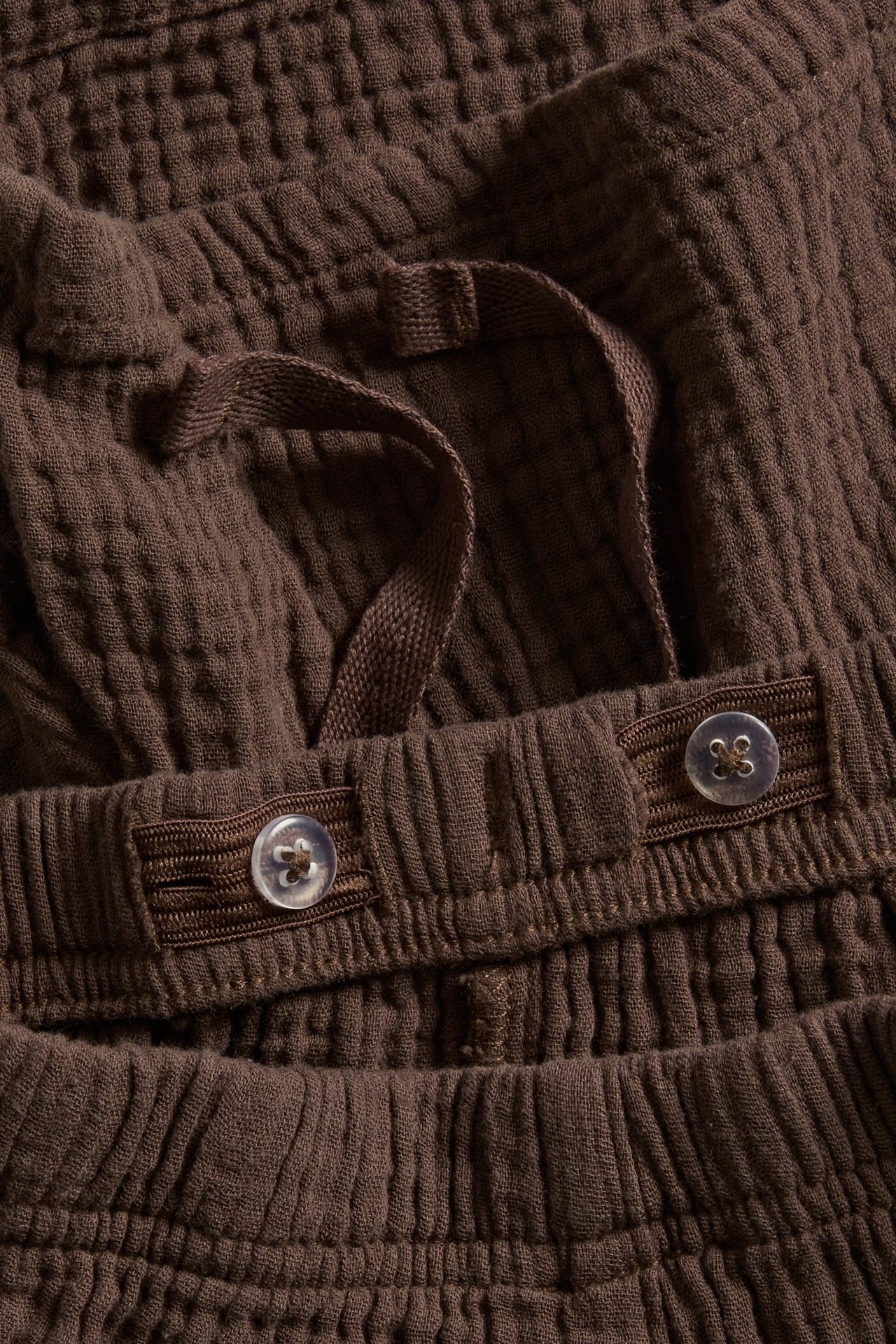 Zbliżenie na brązowe szorty z muślinu, ozdobny sznurek, regulowana gumka z guzikami.