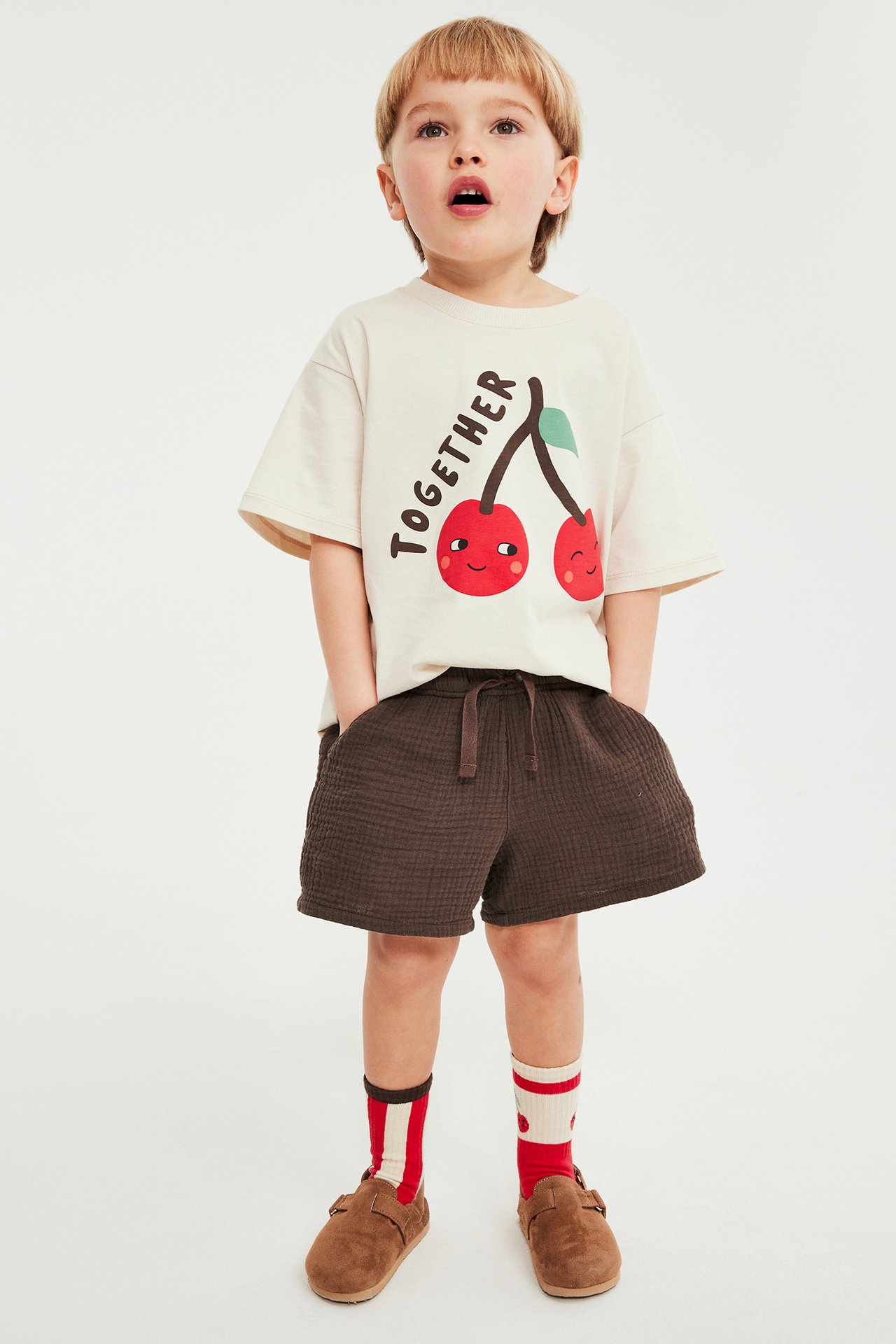 Widok z przodu: Dziecko w brązowych muślinowych szortach, beżowym t-shircie, paskowanych skarpetach i brązowych butach.