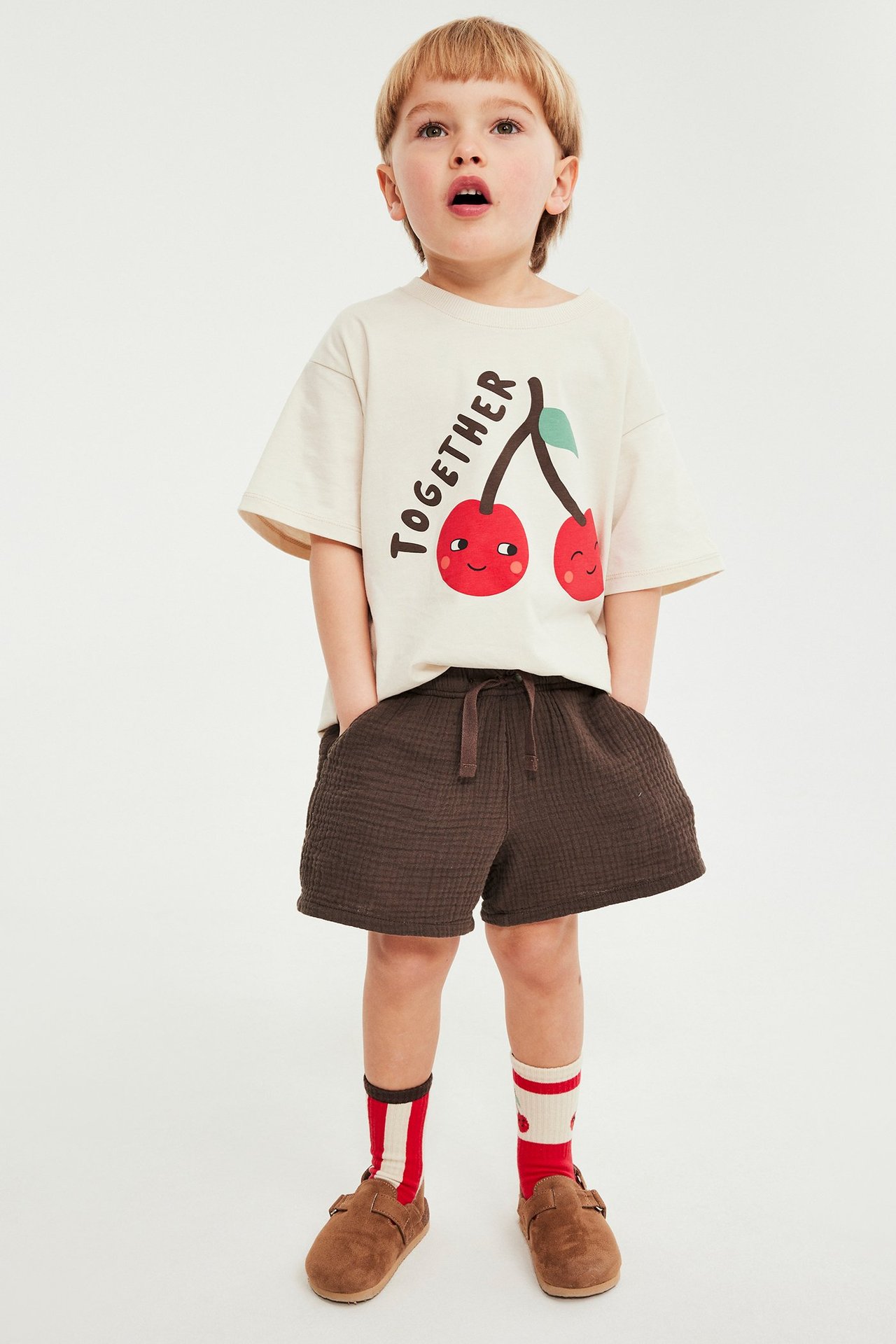 Widok z przodu: Dziecko w brązowych muślinowych szortach, beżowym t-shircie, paskowanych skarpetach i brązowych butach.