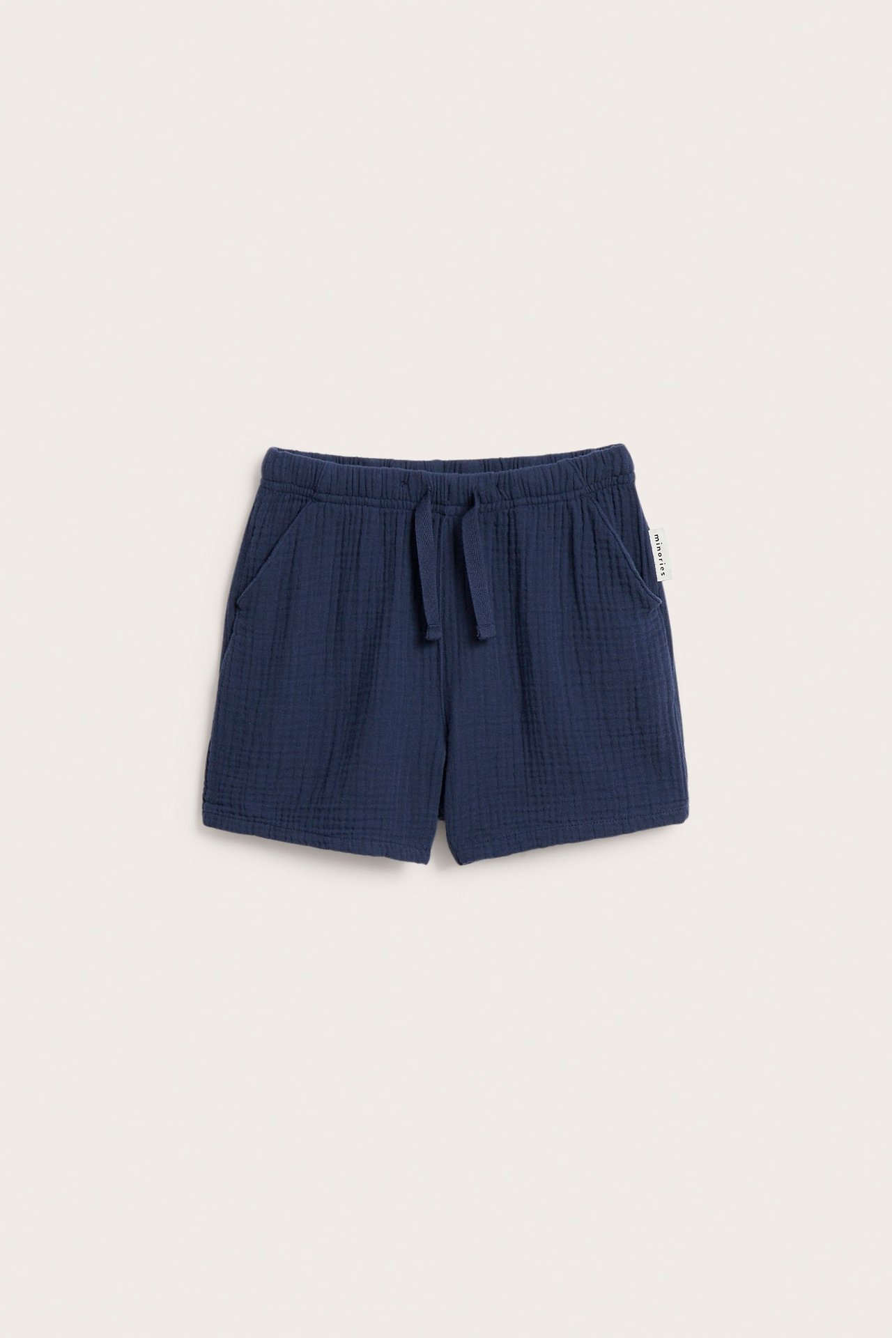 Frontvy av blå muslinshorts för barn med sidfickor och en fejkdragsko i midjan.