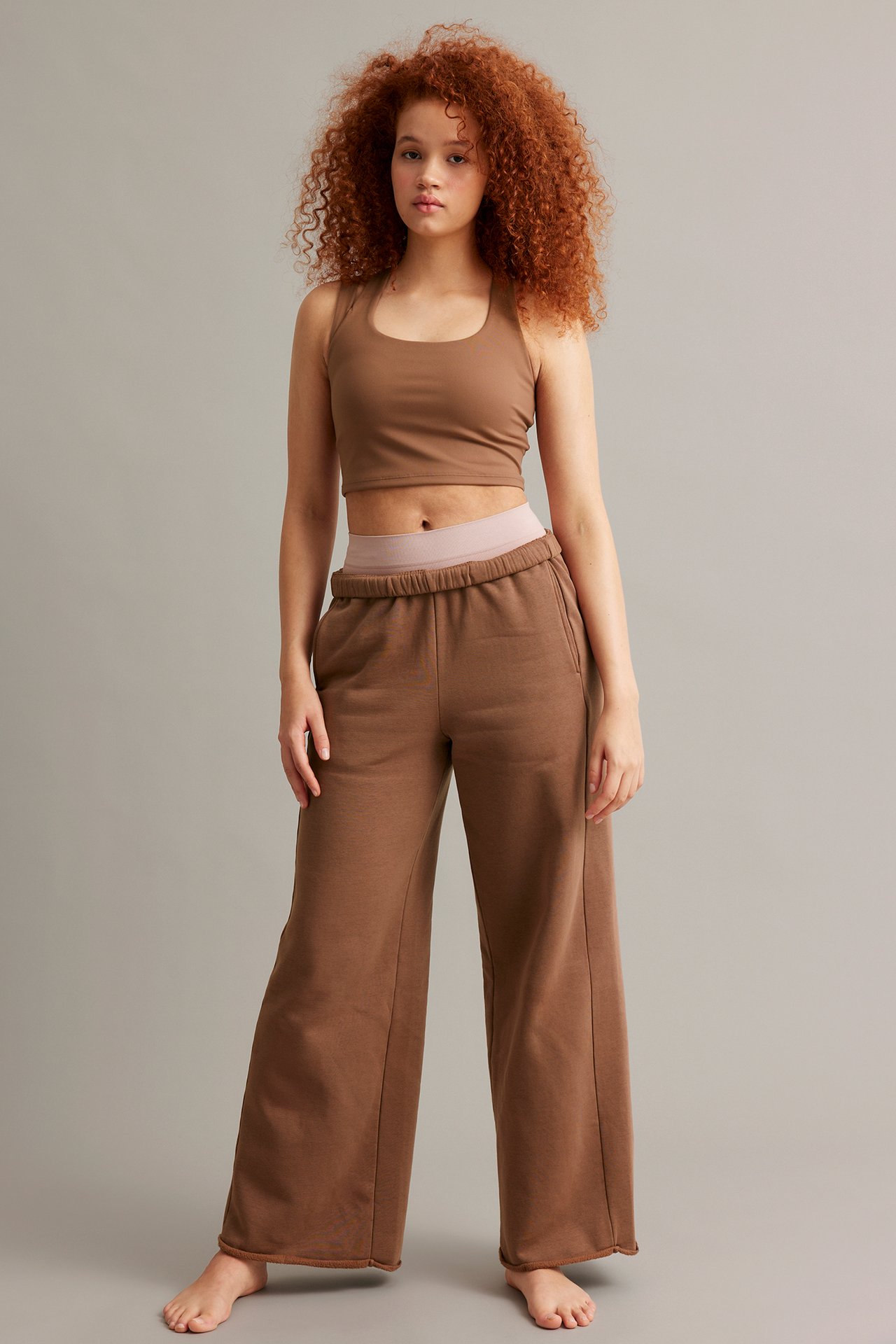 Edestäpäin nainen ruskeassa urheilutopissa ja ruskeissa leveälahkeisissa collegehousuissa, joista näkyy vaaleanpunainen vyötärönauha.