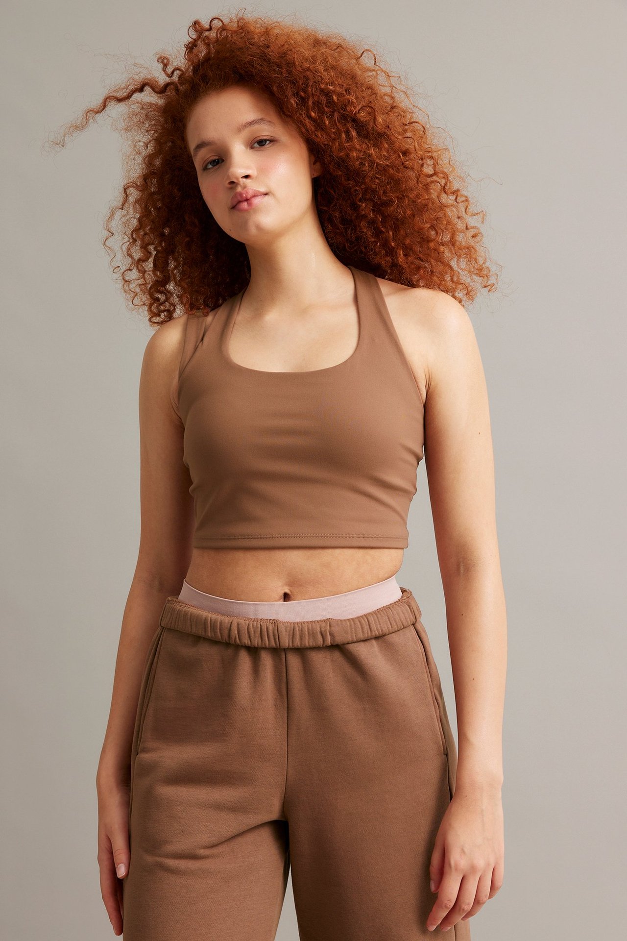 Forfra: kvinne i figurnær brun sportstopp med brede stropper og matchende brune joggebukser.